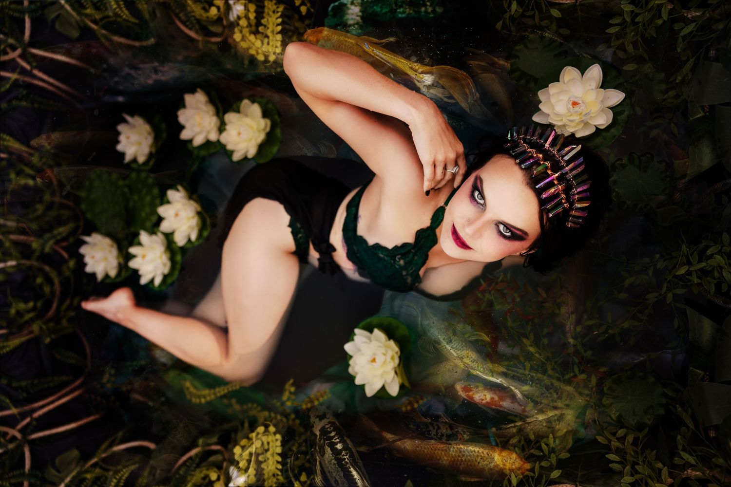 Woman in black swimsuit with crystal crown lying among white water lilies in dark natural pool.