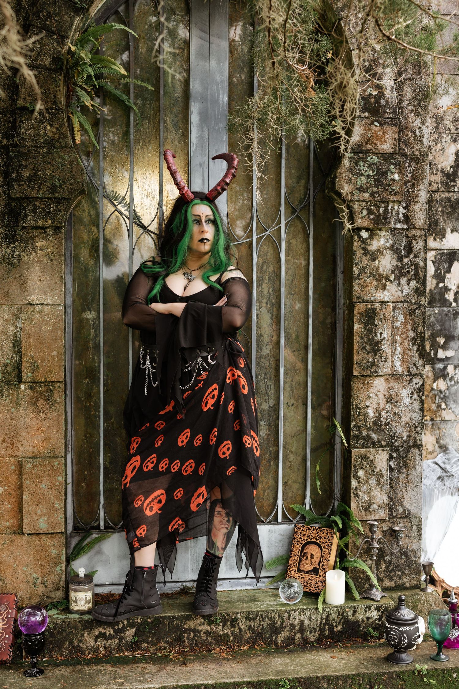 Woman in green hair, horns, and Halloween witch costume poses by mossy stone gate with candles and skull decor.