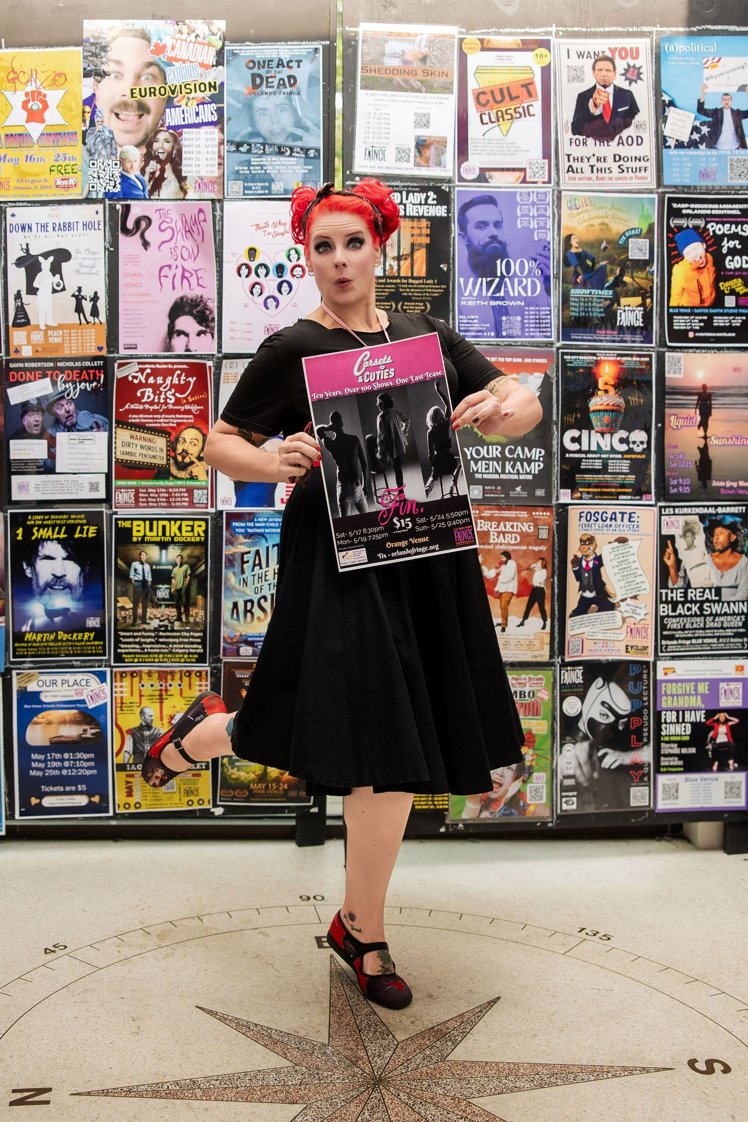 Woman with red hair holding show poster in front of flyer wall.