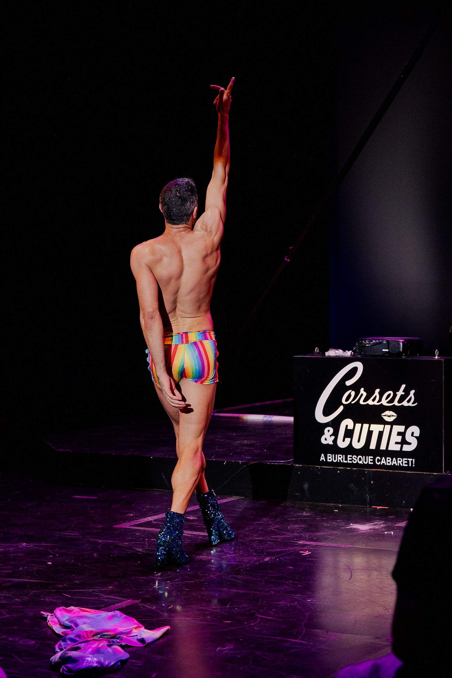 Shirtless male performer in rainbow briefs and glitter heels posing on stage at burlesque cabaret.