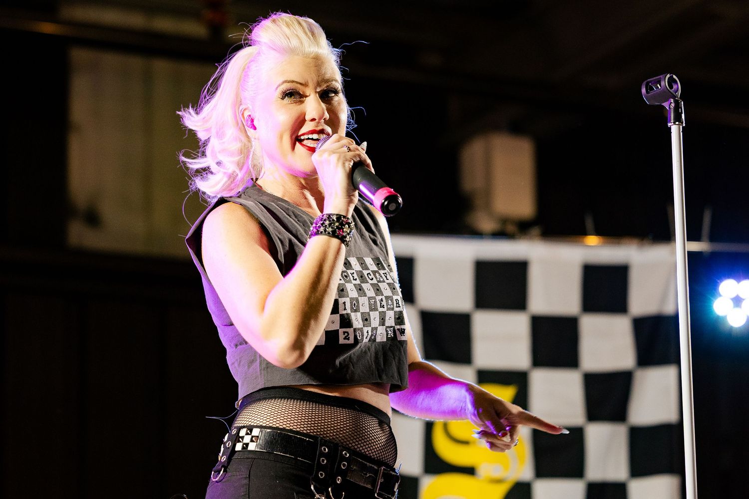 A female singer with pink-blonde hair performs on stage wearing a checkered top with a racing flag backdrop.