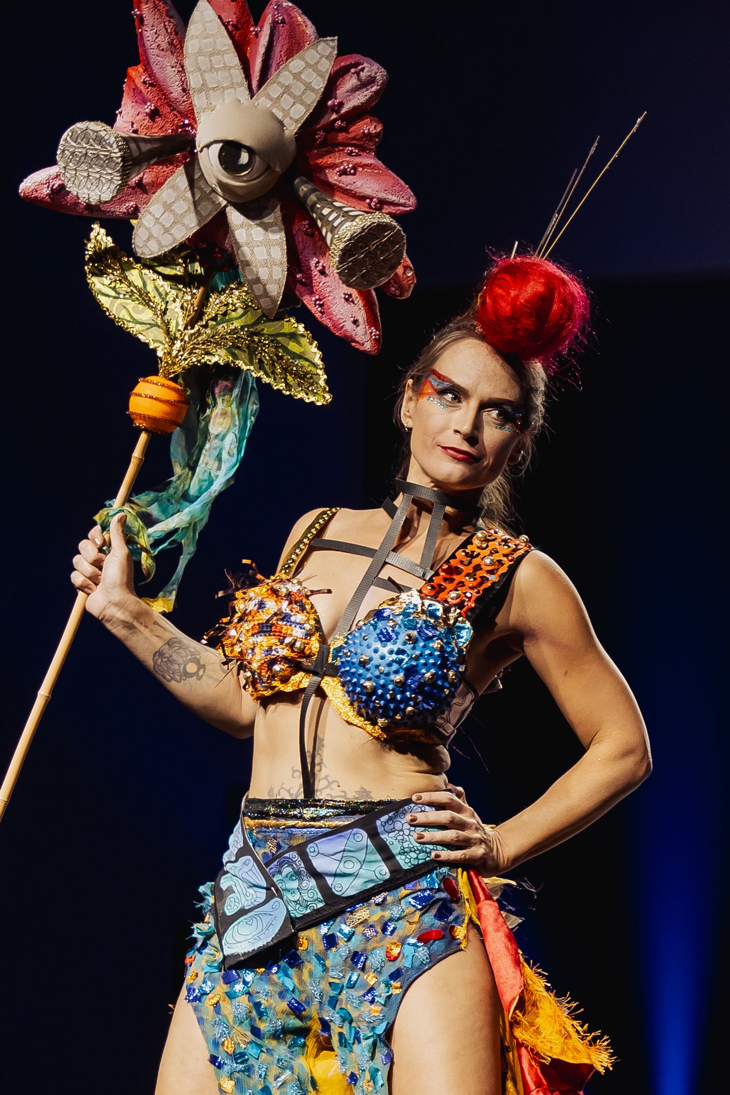 Female model in colorful fantasy costume holding a large flower prop on runway.