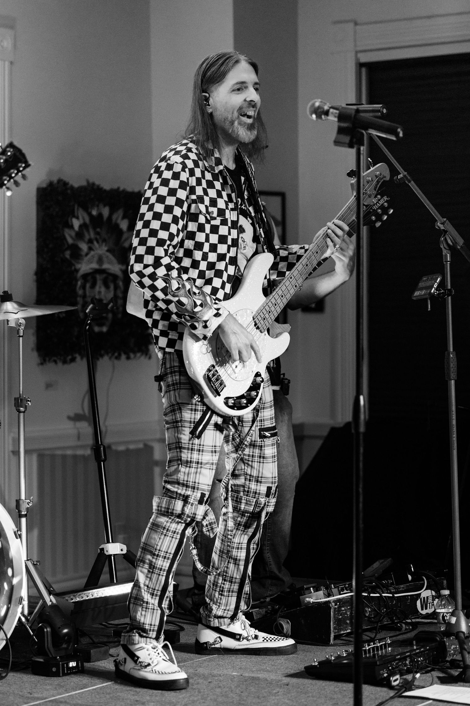 Black and white photo of a long-haired guitarist in checkered shirt and plaid pants performing on stage with electric guitar.