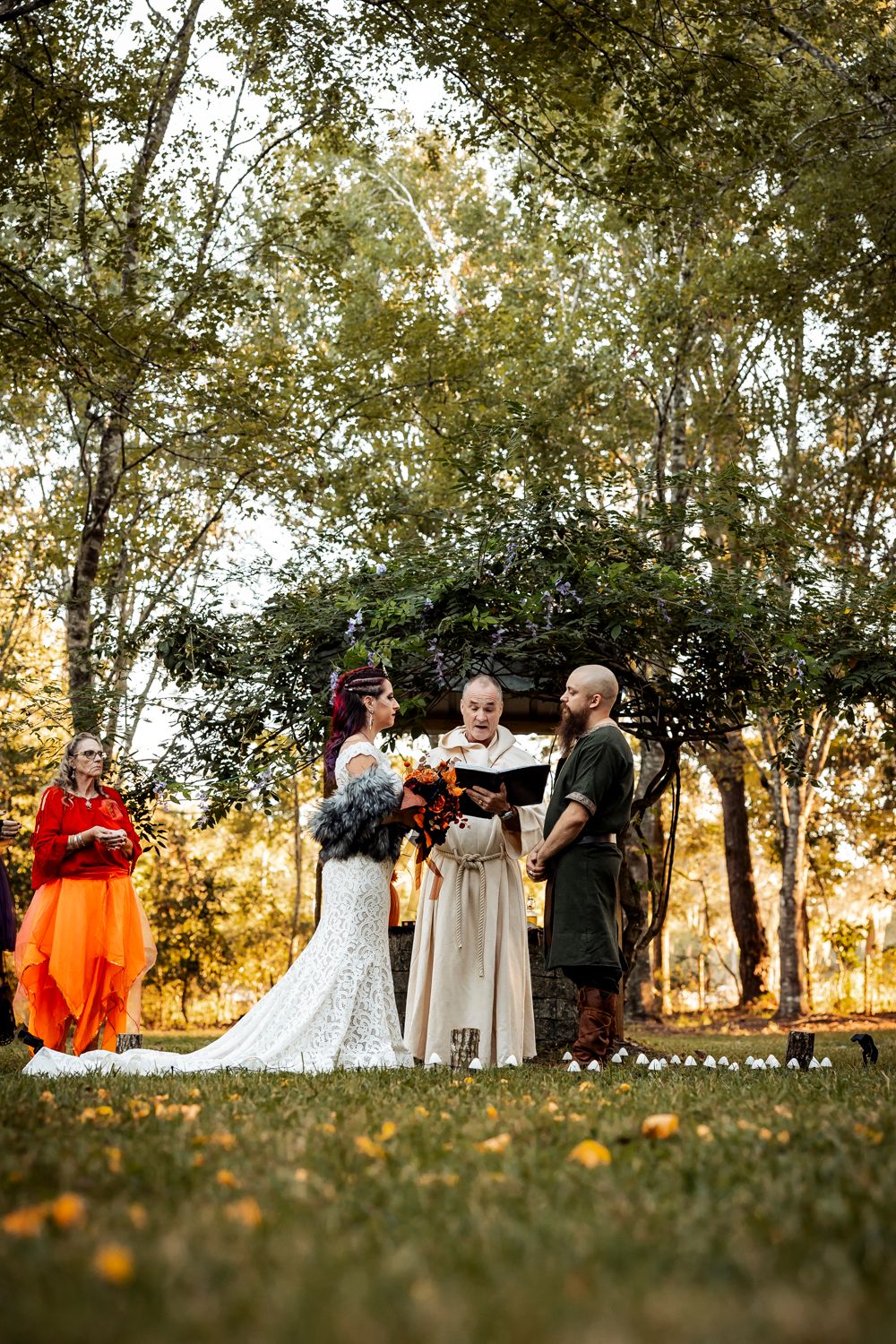 Viking-themed outdoor wedding ceremony with bride, groom, and officiant under floral arch at sunset.