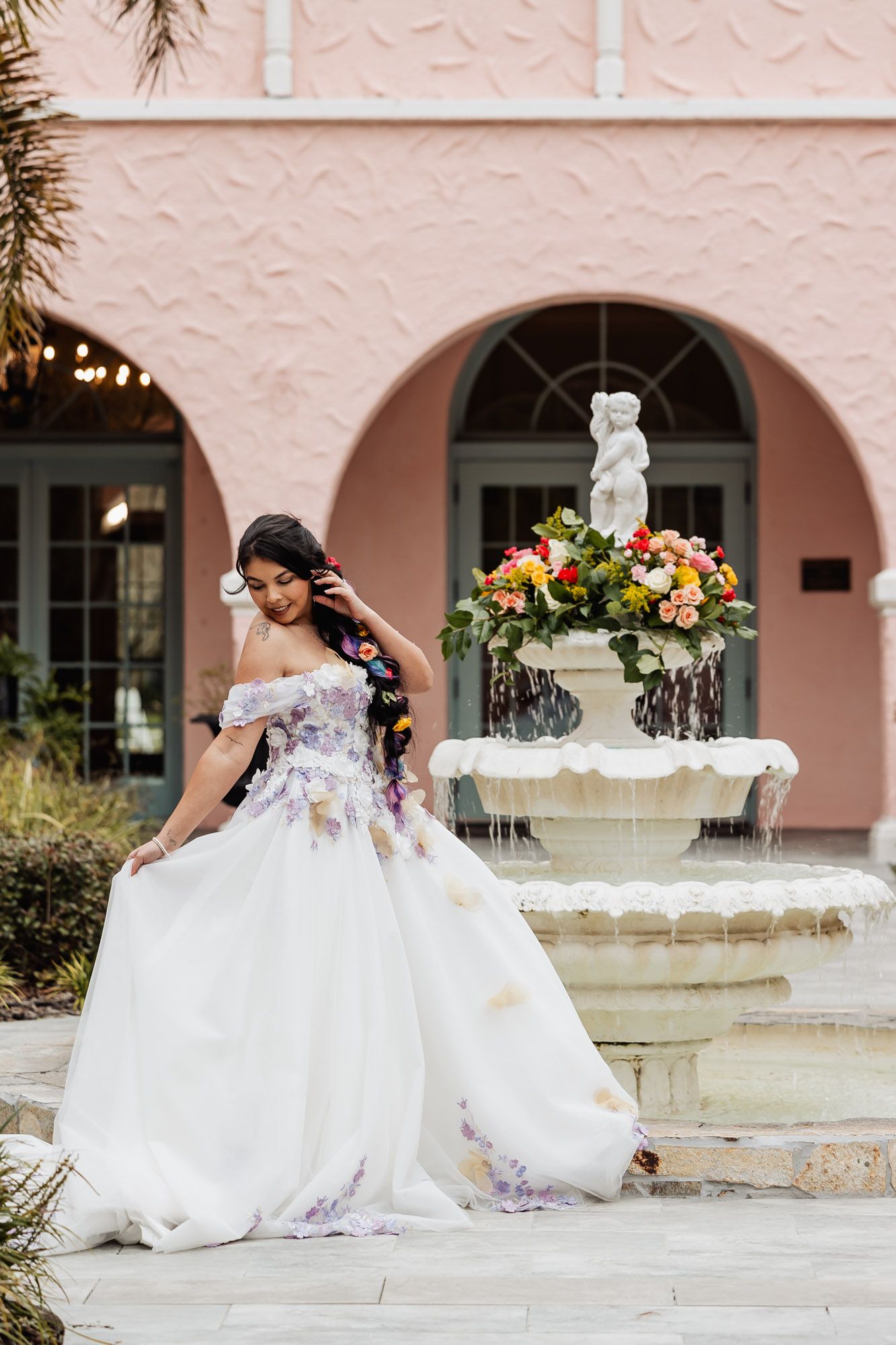 Bride in floral off-shoulder ball gown twirling near ornate fountain at pink Mediterranean venue.
