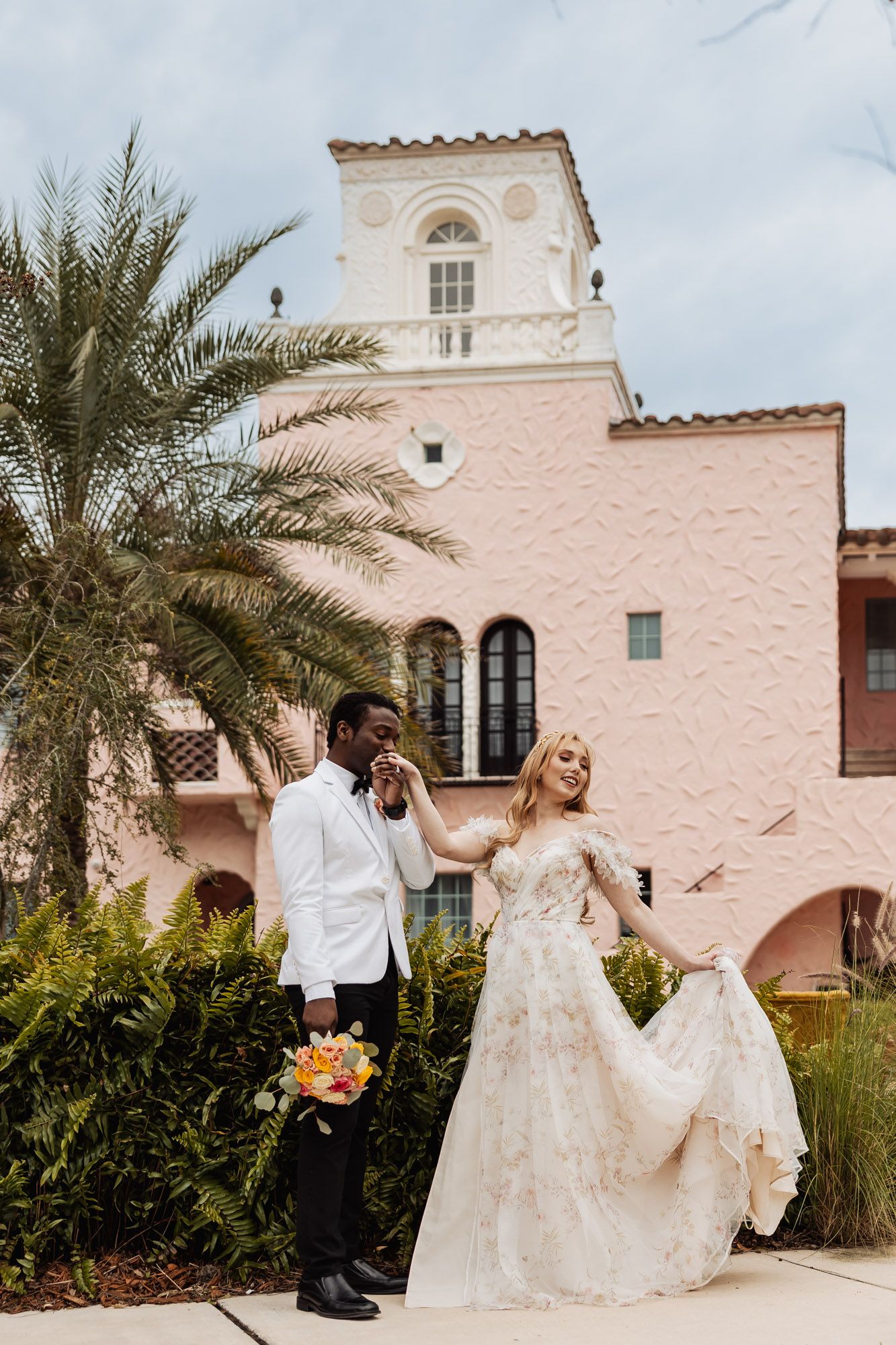 Interracial wedding couple walking hand-in-hand outside pink Spanish colonial building with palms.