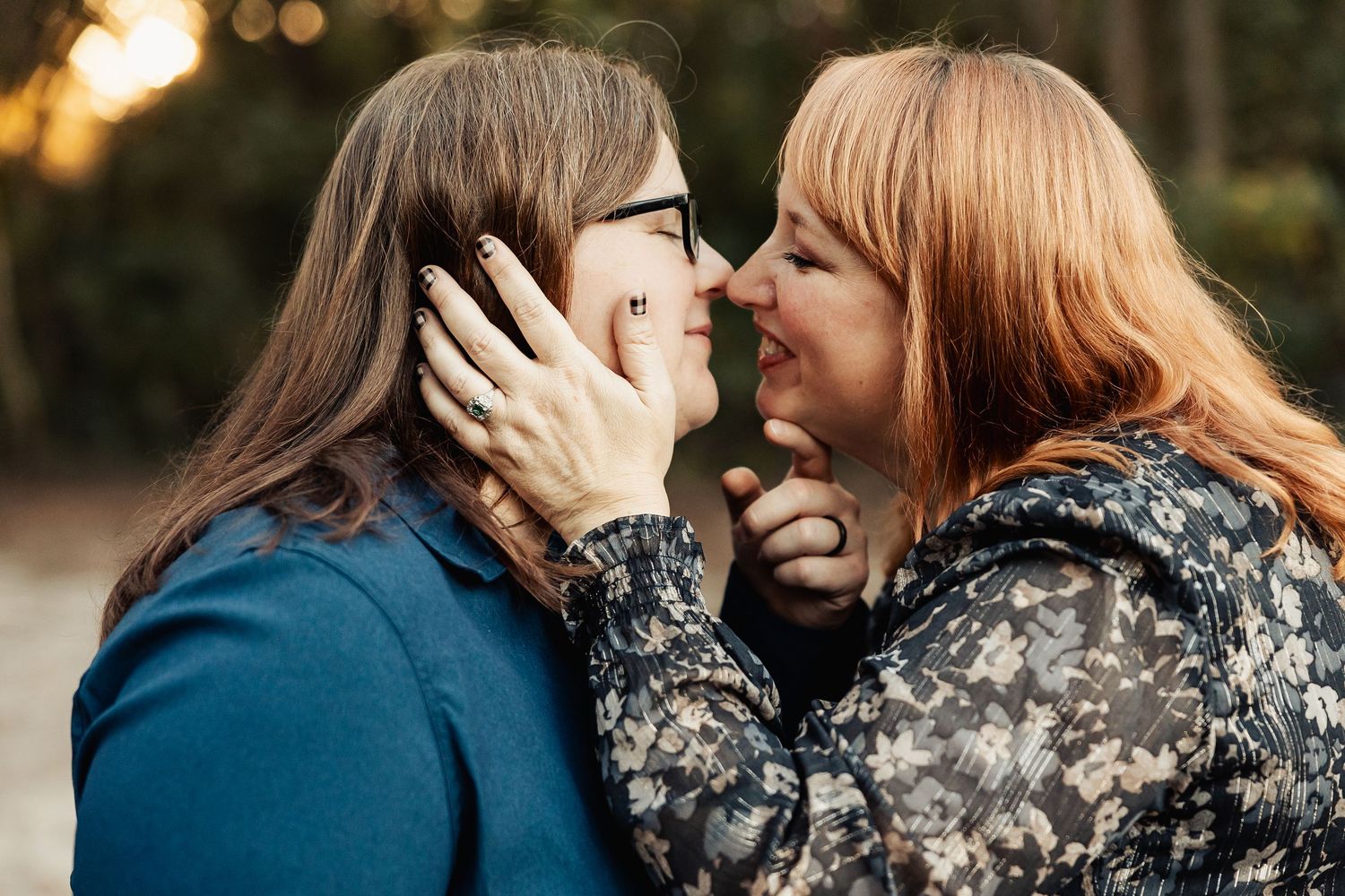 Two women share a tender nose-to-nose moment, hands cupping each other's faces during a romantic outdoor photoshoot.