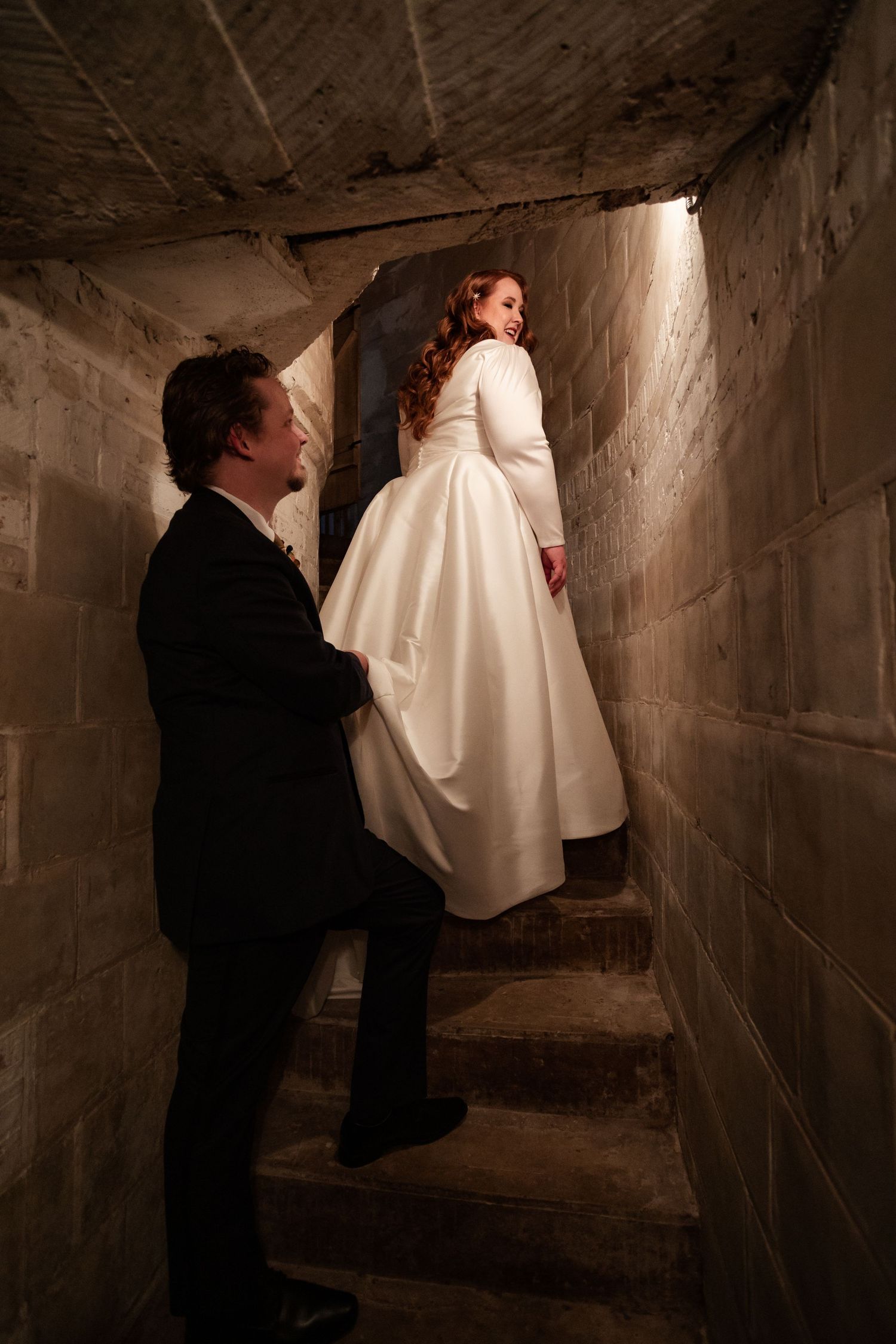 Bride in elegant white gown leads groom up stone staircase in dimly lit historic venue.