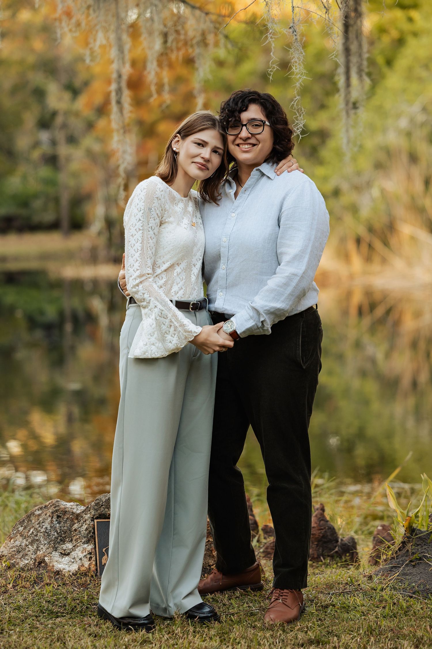 Two women pose together smiling by a tranquil pond surrounded by autumn trees in soft natural light.