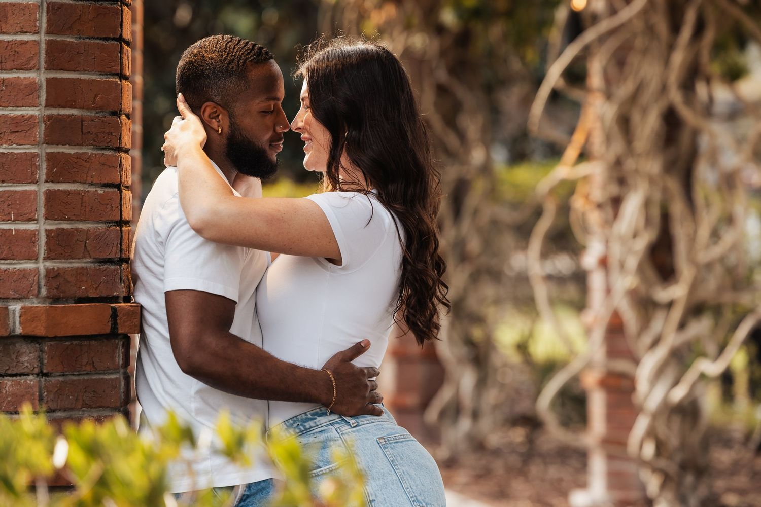 A romantic couple in casual white shirts and jeans embraces outdoors against a brick wall in warm sunlight.