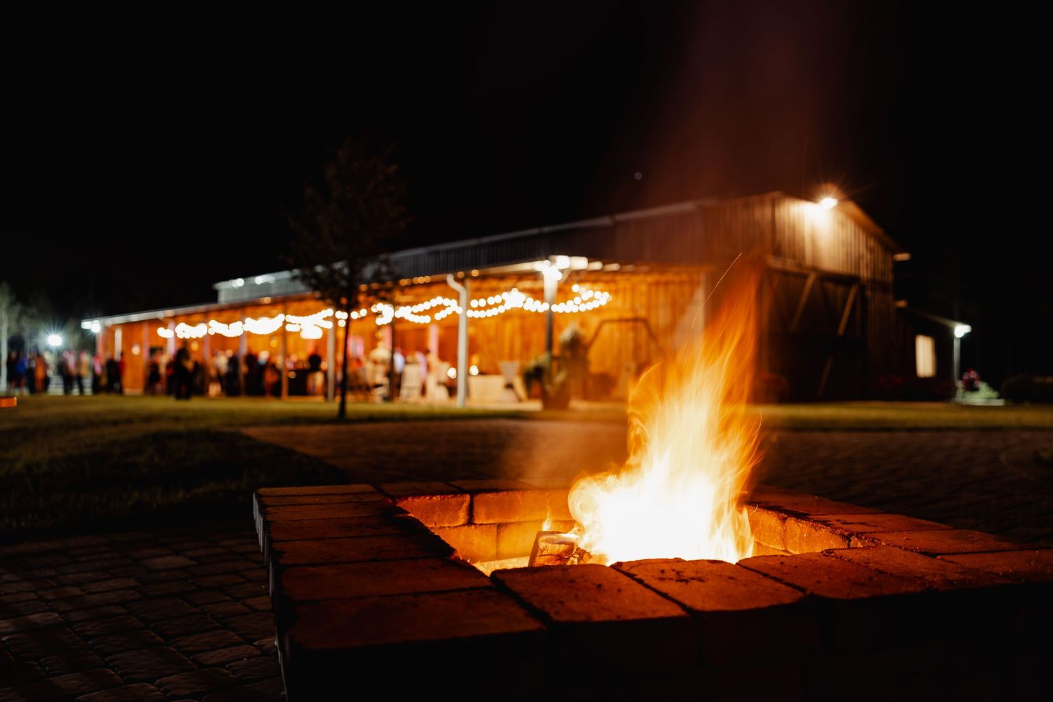 Large bonfire burning outside illuminated barn wedding venue at night.