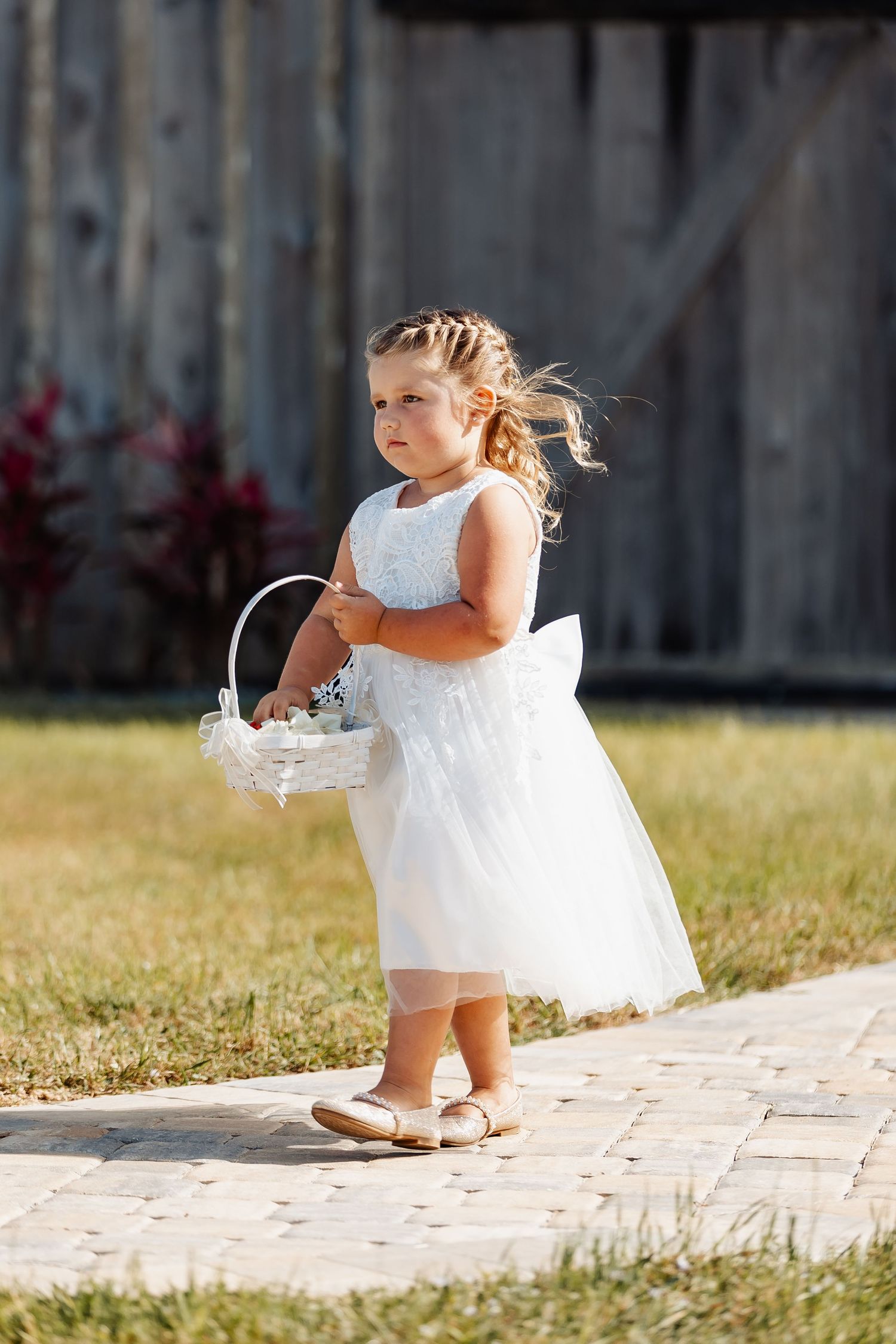 A flower girl in white dress carries a basket along a stone path.