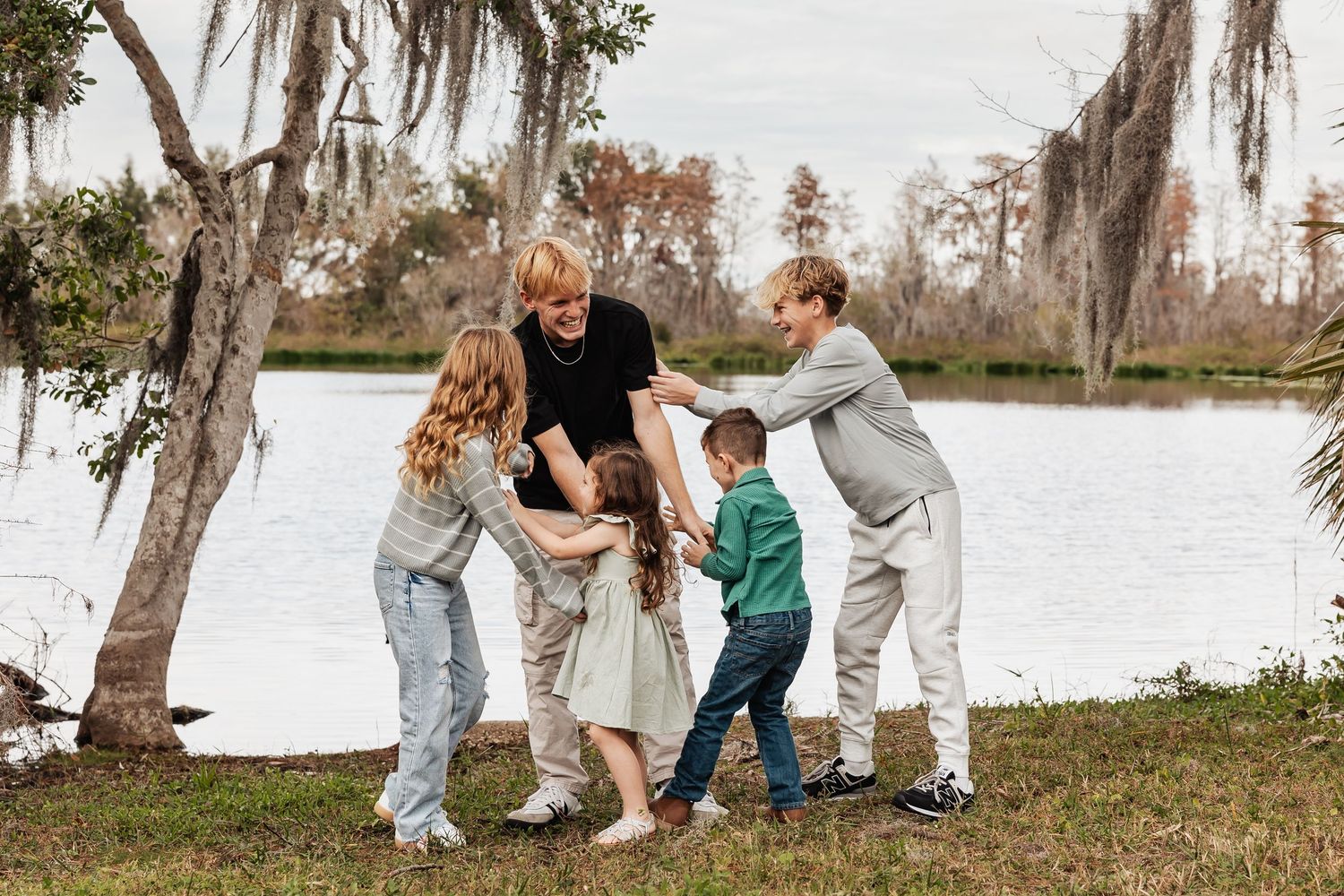 A father and three children playfully interact together on a grassy lakeside with moss-covered trees.