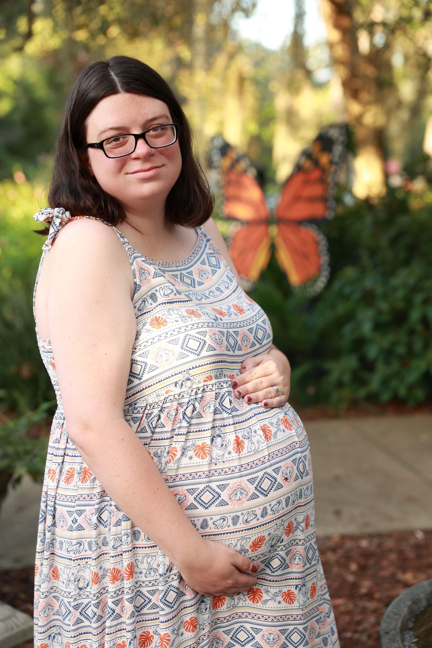 A smiling pregnant woman in a patterned dress poses outdoors near decorative butterfly wings.