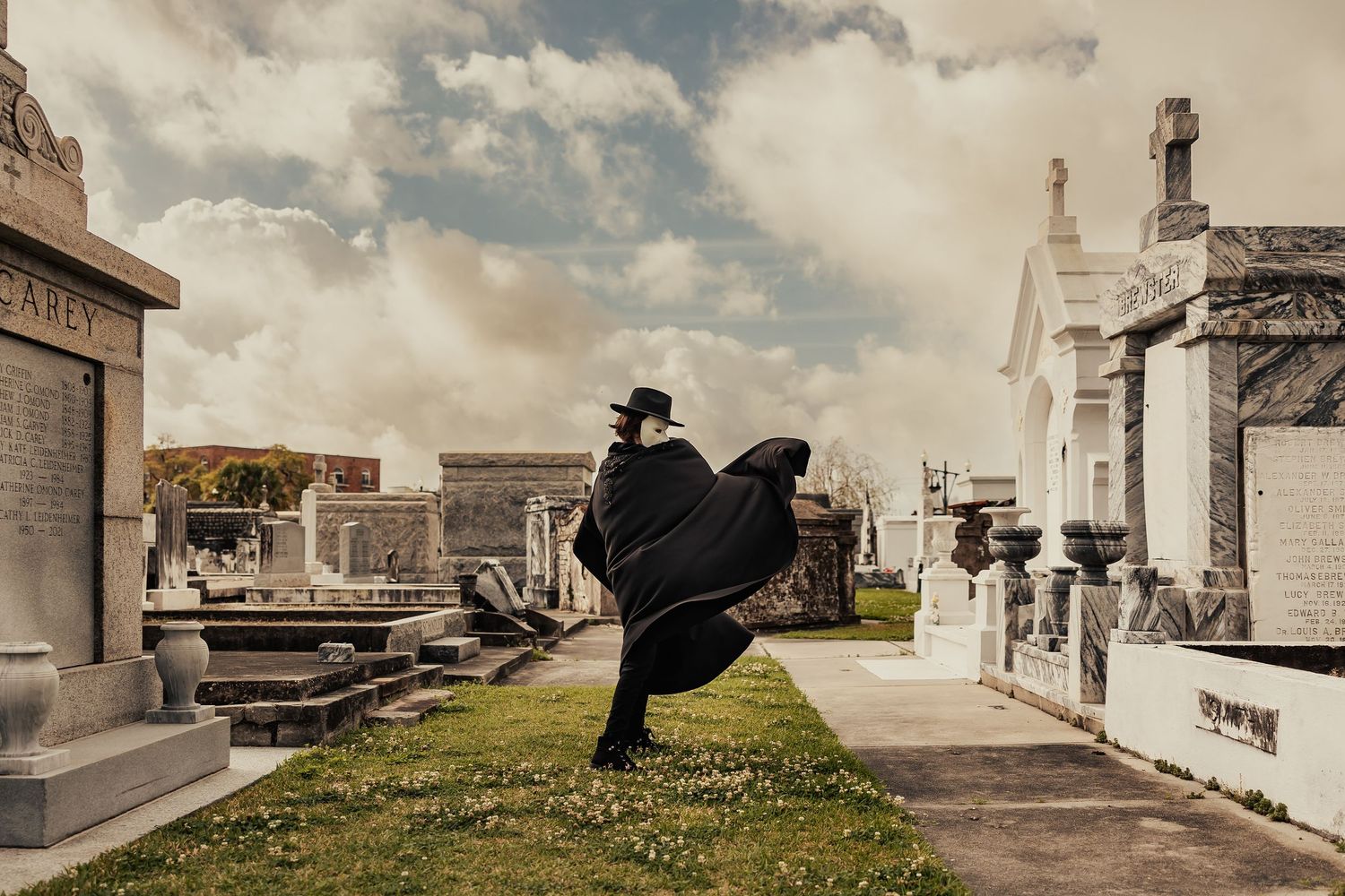 Mysterious caped figure in black hat walking through historic above-ground cemetery on cloudy day.