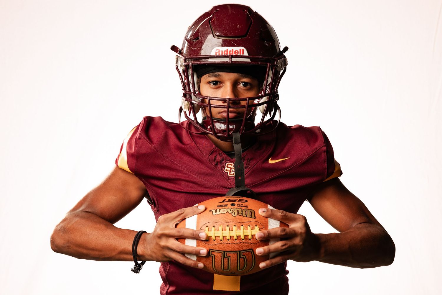 Young football player in maroon Nike uniform holding a football, posing for a professional sports portrait photo.