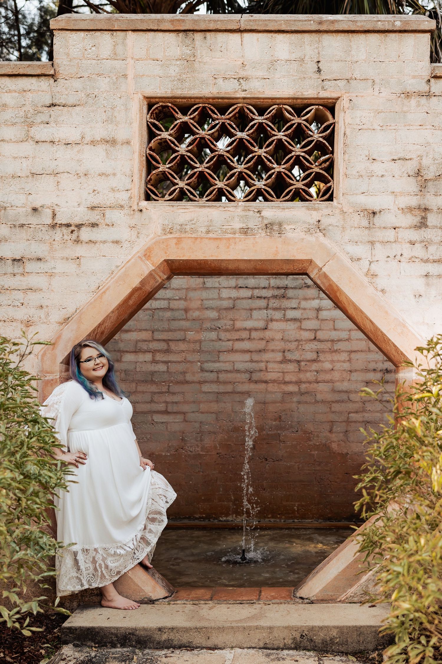 Pregnant woman in white dress sitting by a brick fountain with hexagonal arch in a garden setting.
