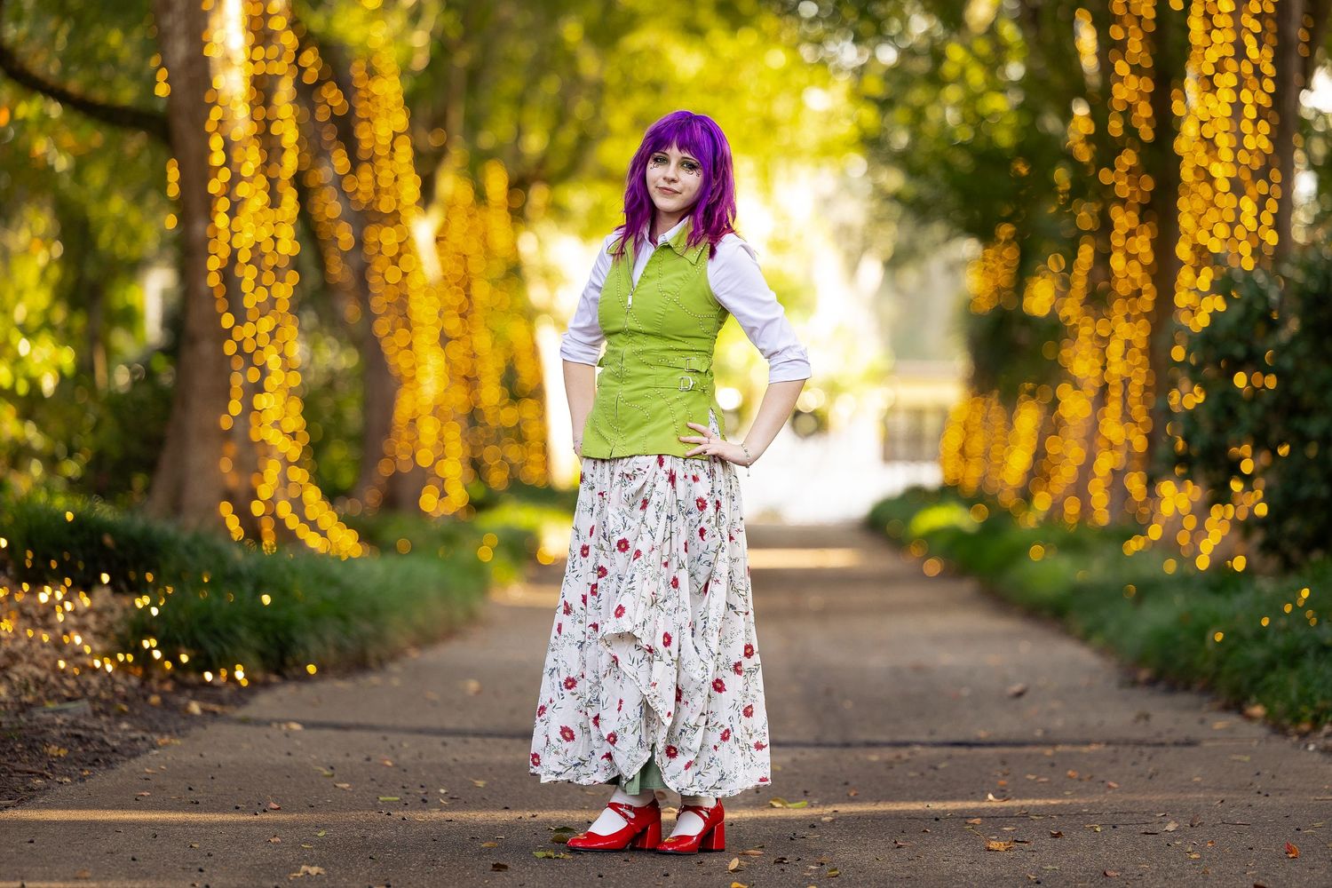 Woman with purple hair in green vest, floral skirt, and red shoes stands on a tree-lined path in golden light.