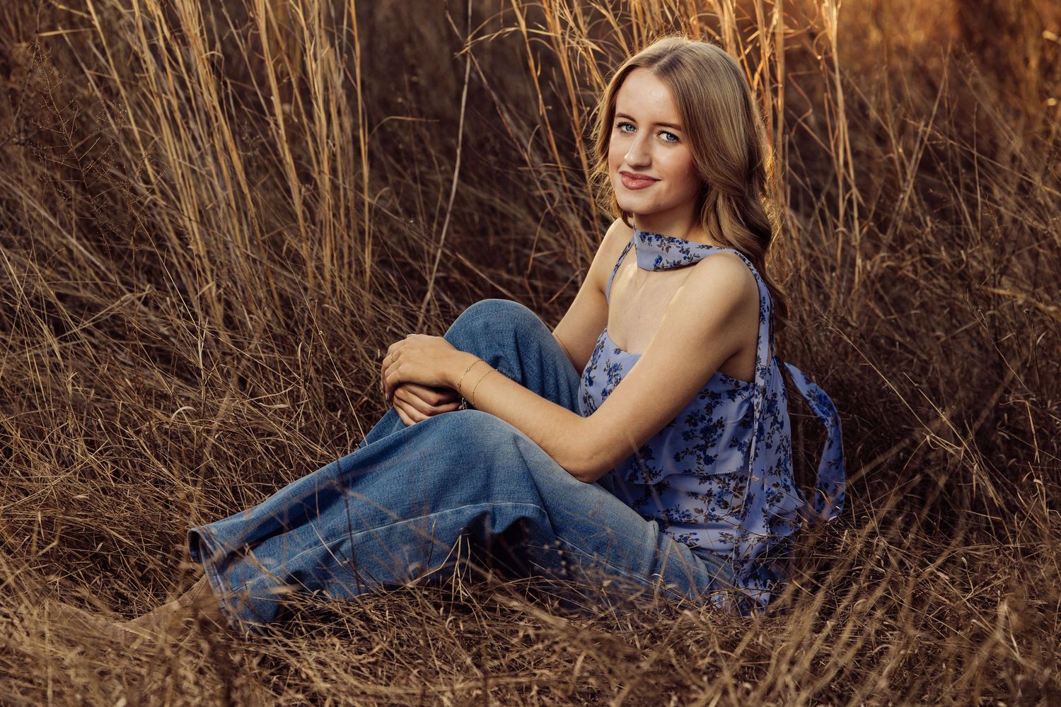 Young woman in blue floral top and jeans sitting in golden tall grass field during sunset.