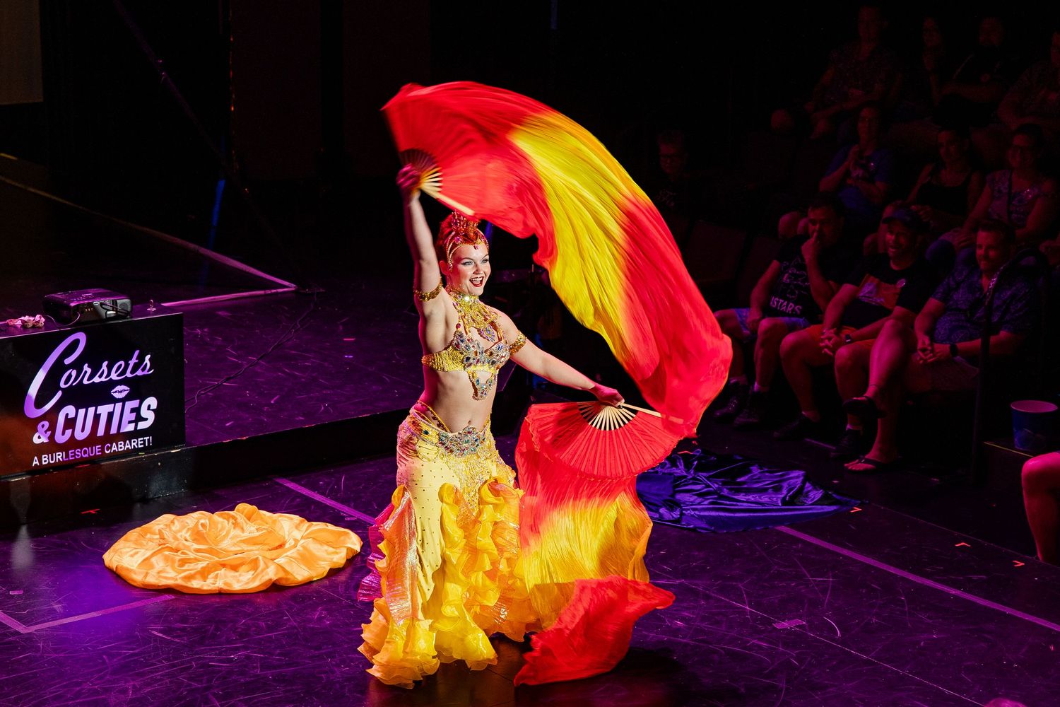 Burlesque dancer in yellow and red costume waving large colorful fans on stage.