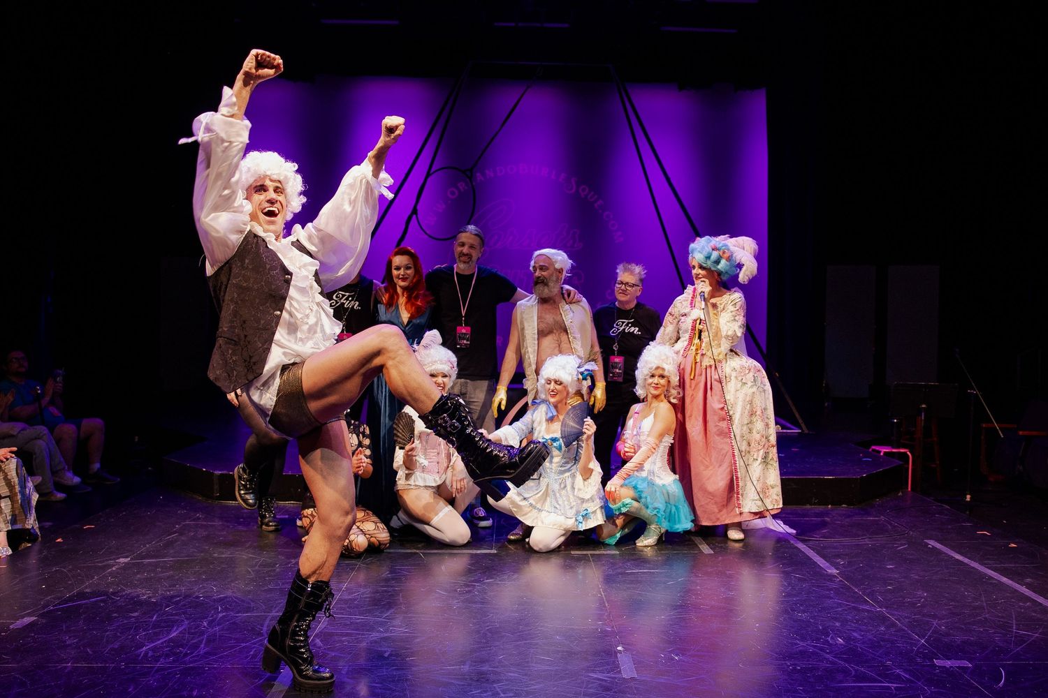 Burlesque troupe in rococo costumes and white wigs take a lively bow on stage under purple lights.