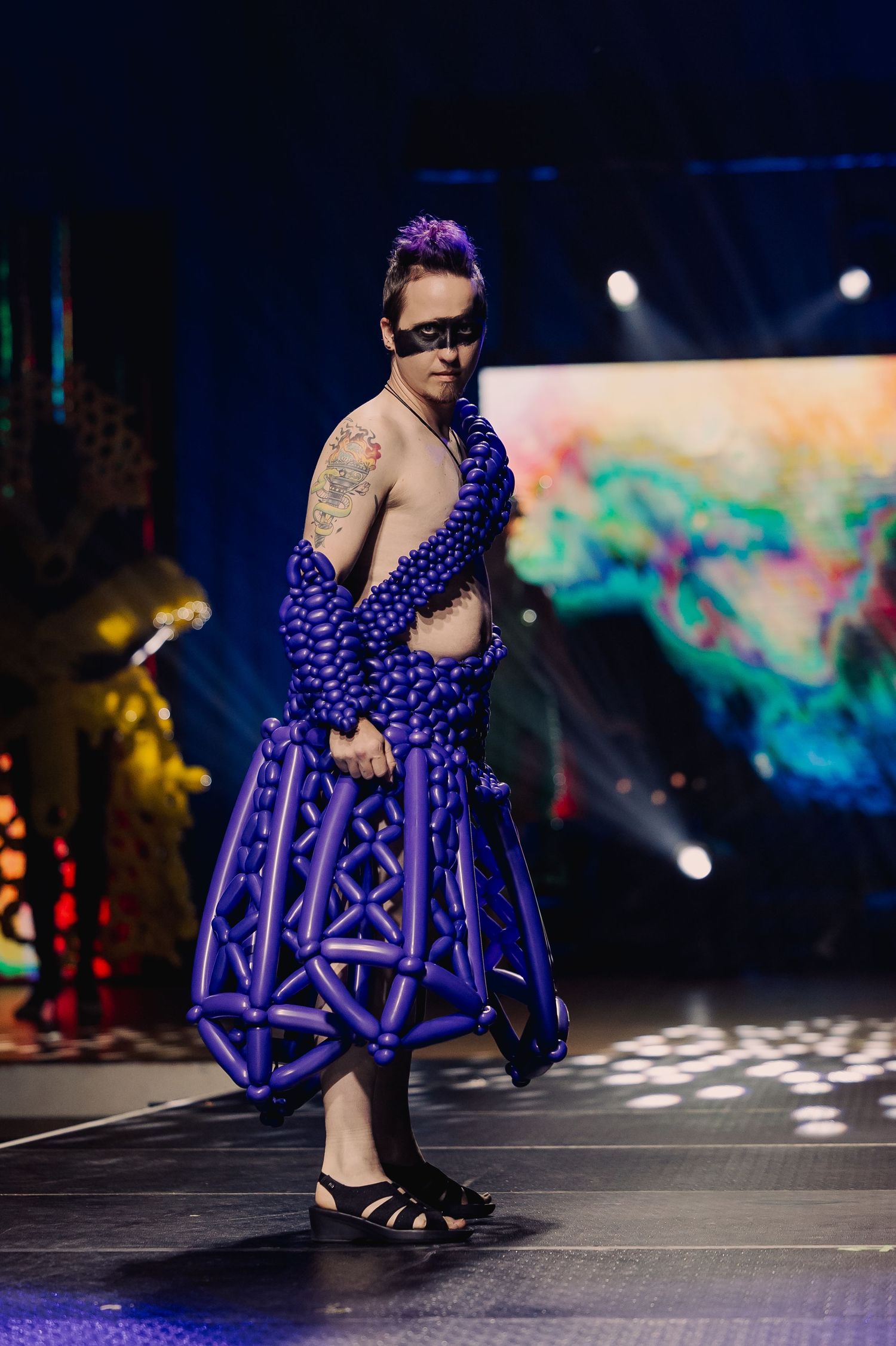 Model wearing a purple balloon skirt and jacket with mask walks the runway at a fashion show.