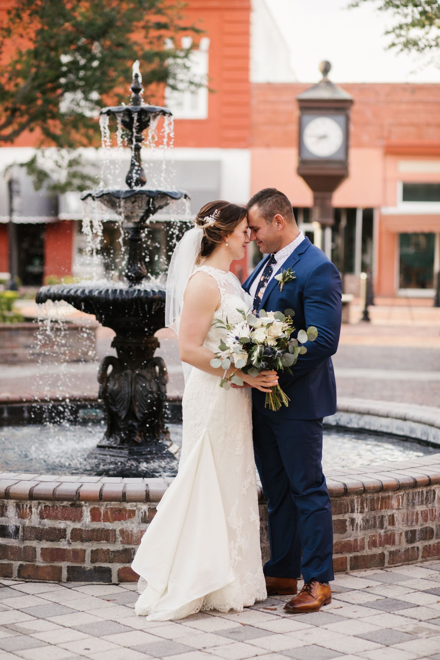 Bride and groom share a romantic moment by a black tiered fountain, she holds white bouquet, he wears navy suit.