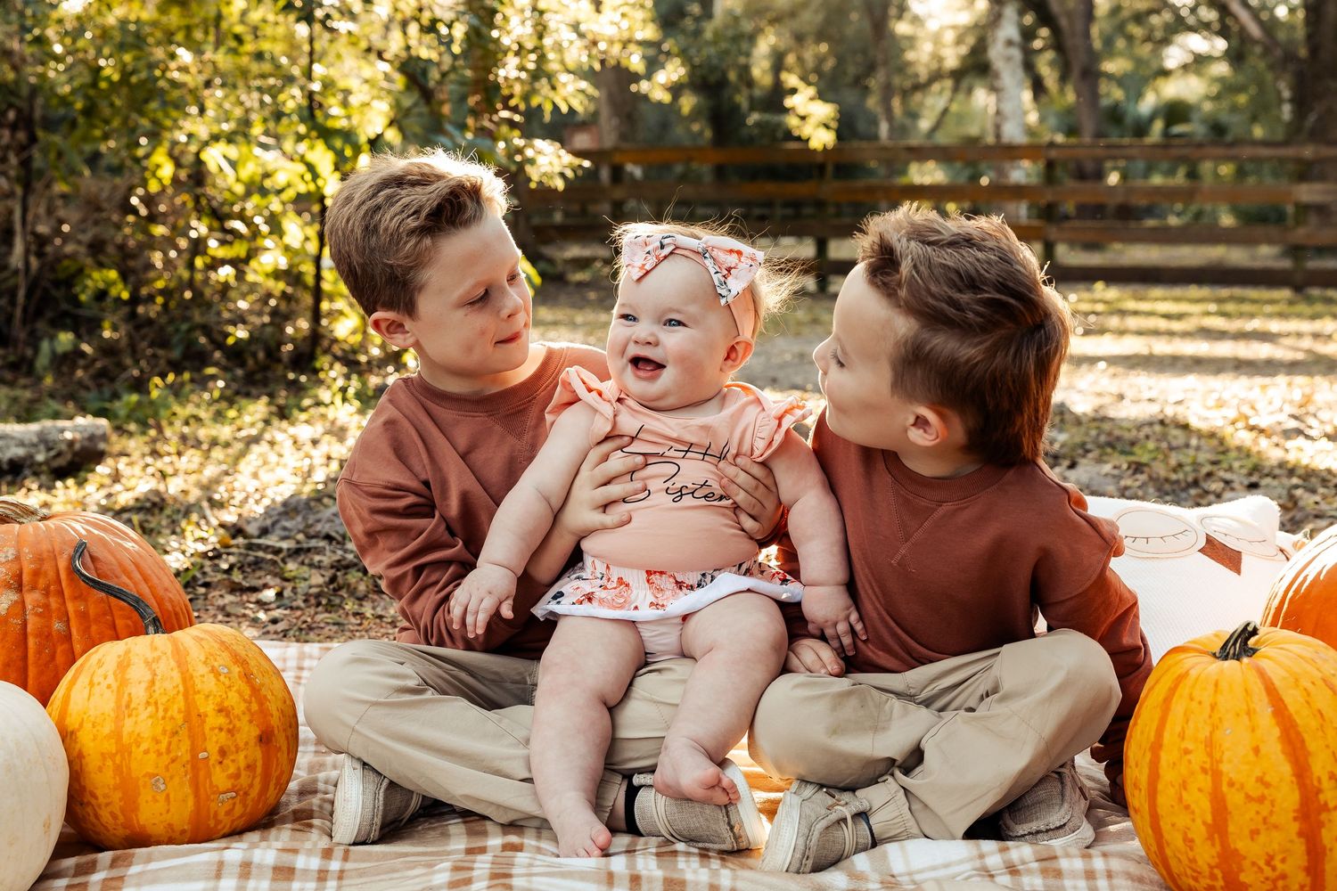 Two brothers sitting on a blanket with their baby sister surrounded by orange pumpkins outdoors.
