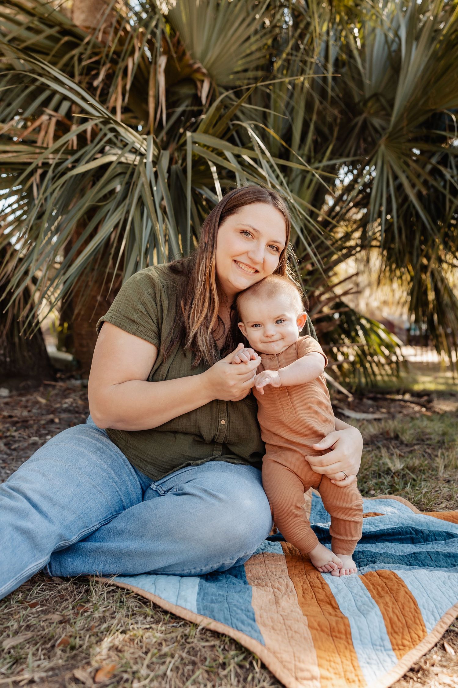 Smiling mother sitting on a blanket outdoors with her baby in front of palm trees.