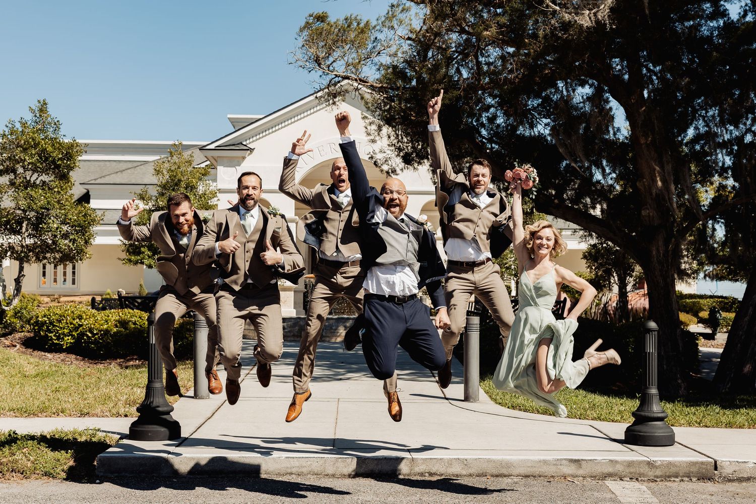 Groom in navy suit jumps with excited groomsmen in tan suits and bridesmaid outside wedding venue.