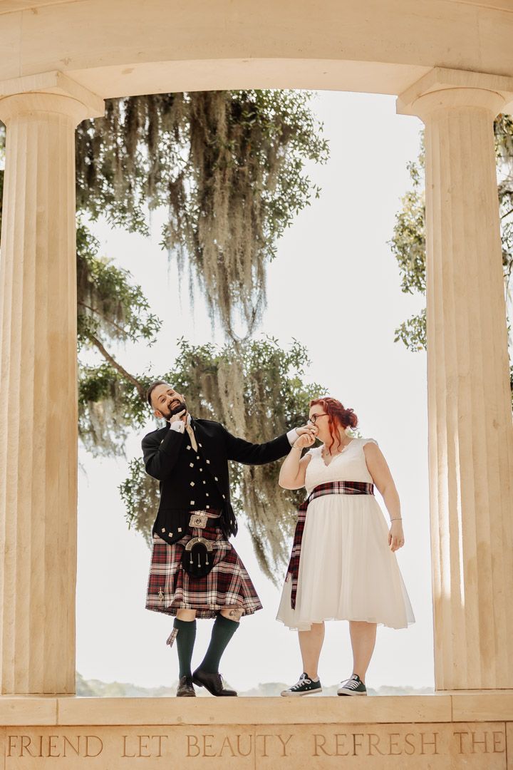 Groom in tartan kilt playfully feeding bride under a columned pavilion with Spanish moss in background.
