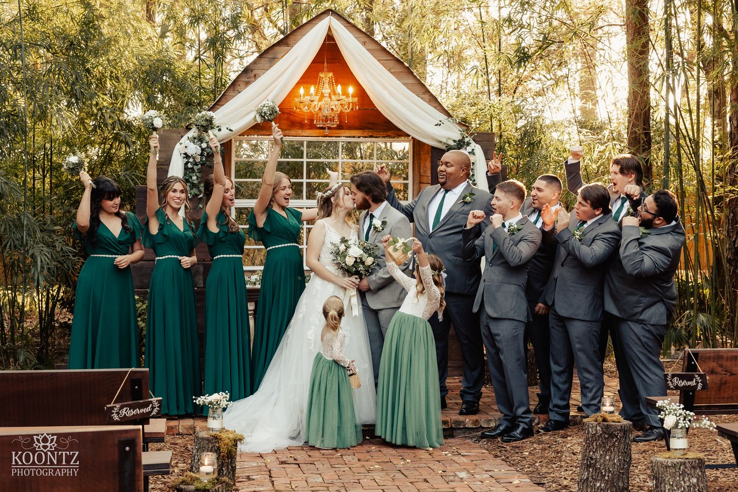 Wedding party celebrates with bride and groom at rustic woodland altar, bridesmaids in emerald green gowns.