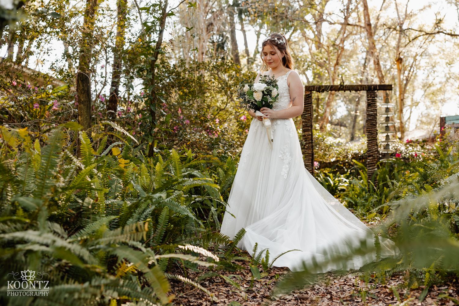 Bride in a flowing white gown stands among green ferns in a sunlit woodland garden setting.