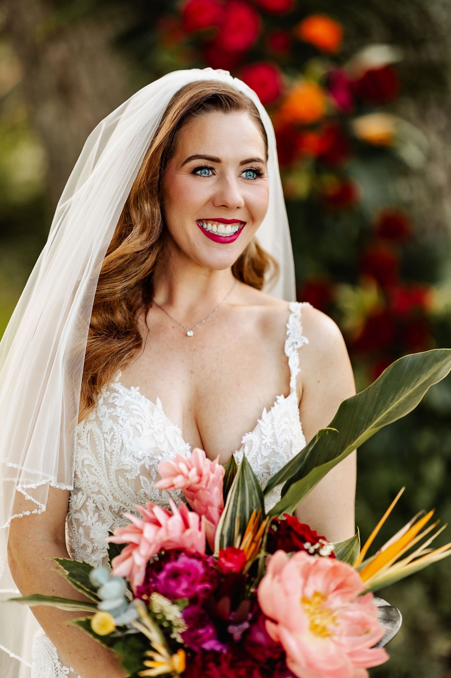 Radiant bride with blue eyes and red lips holds vibrant tropical bouquet with colorful flowers.