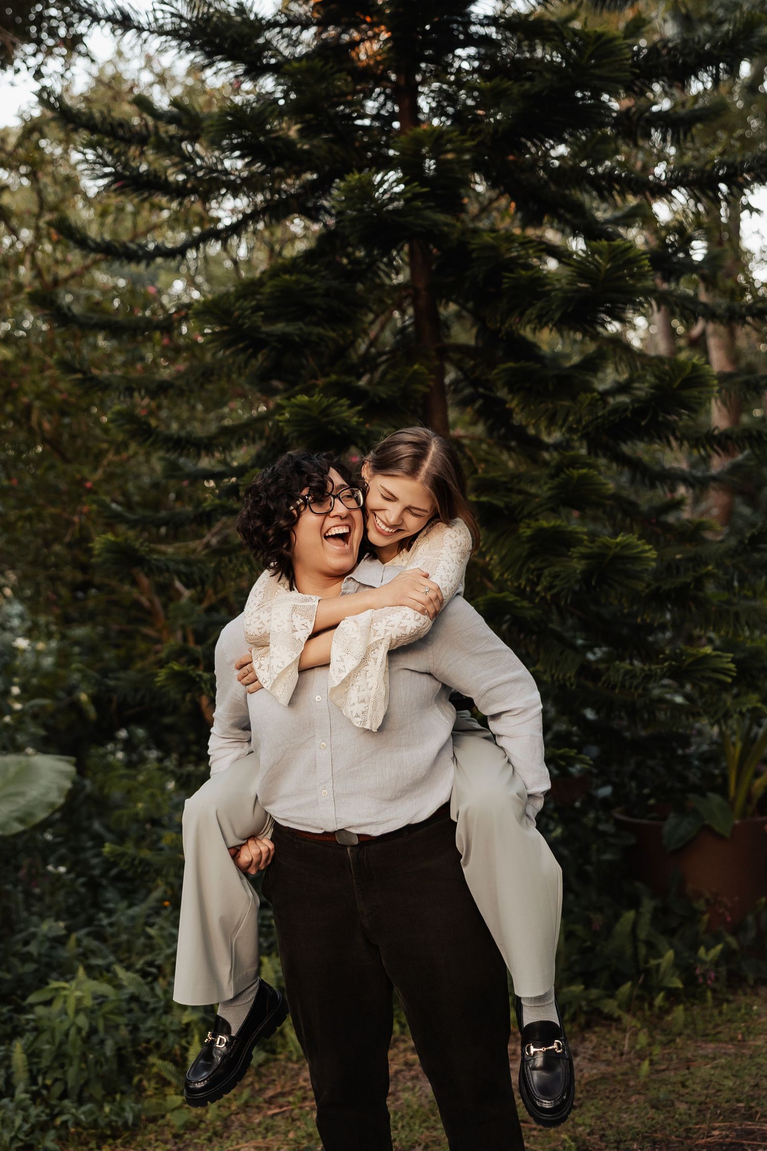 A woman giving another woman a piggyback ride, both laughing in front of evergreen trees.