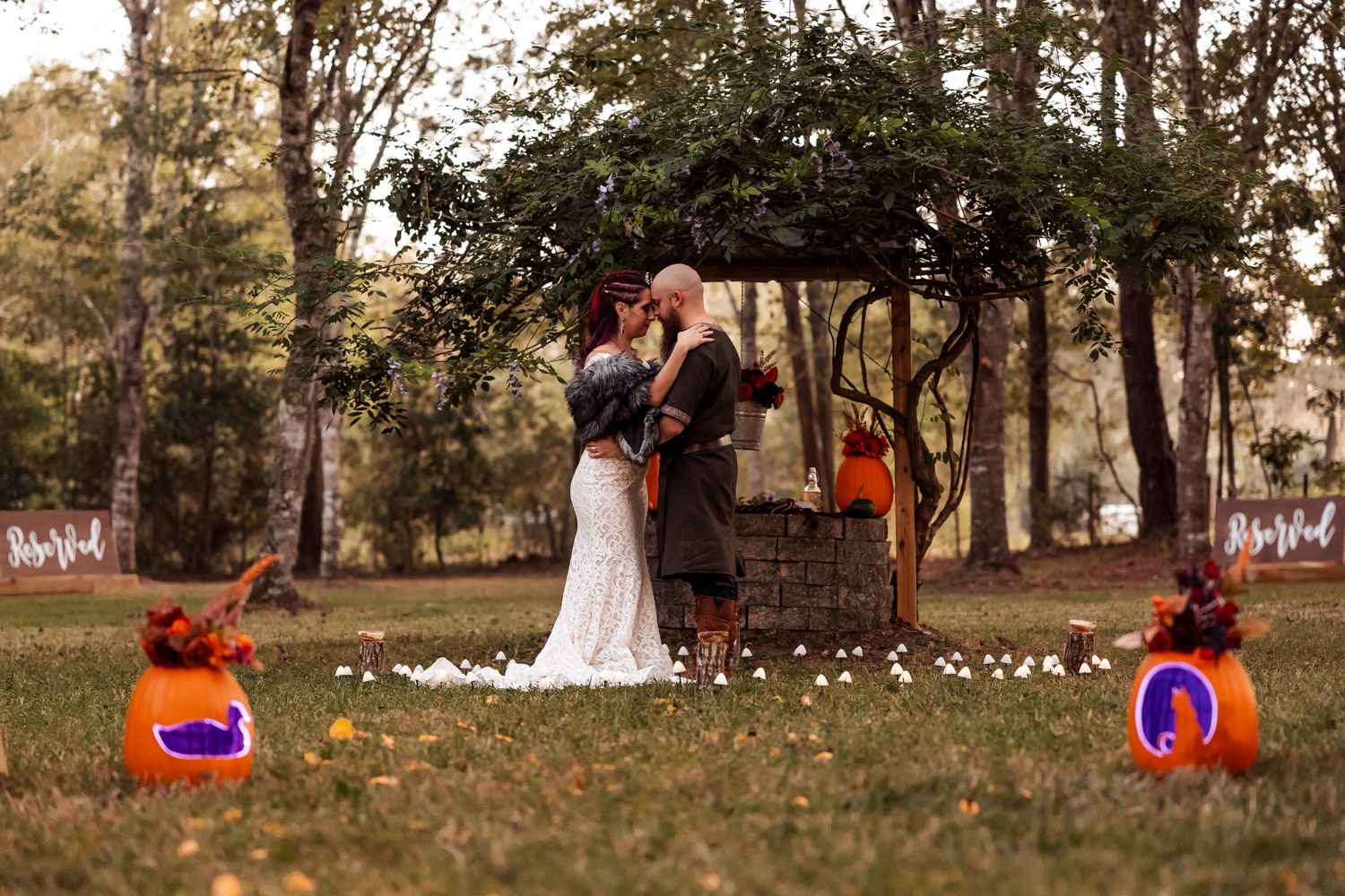 Viking wedding couple kisses at sunset under rustic arch flanked by glowing carved pumpkins outdoors.