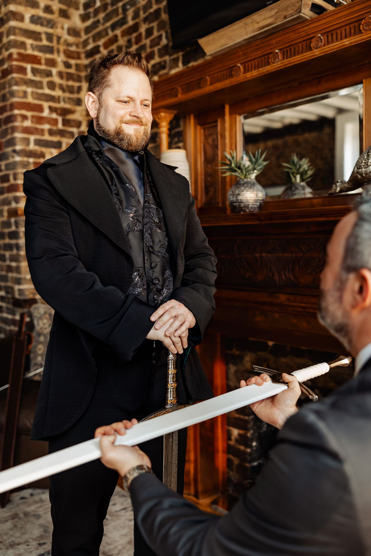 Bearded groom in black suit receiving a sword from groomsman near ornate fireplace.