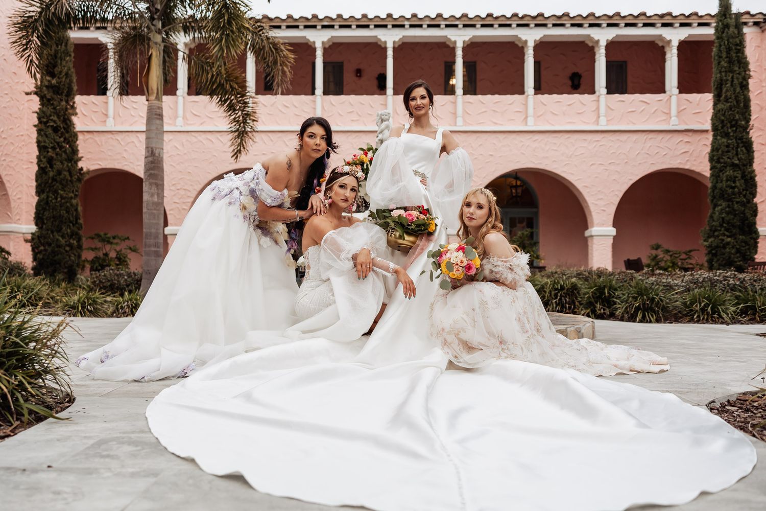 Four brides in white wedding dresses pose together outside a pink Spanish-style building.