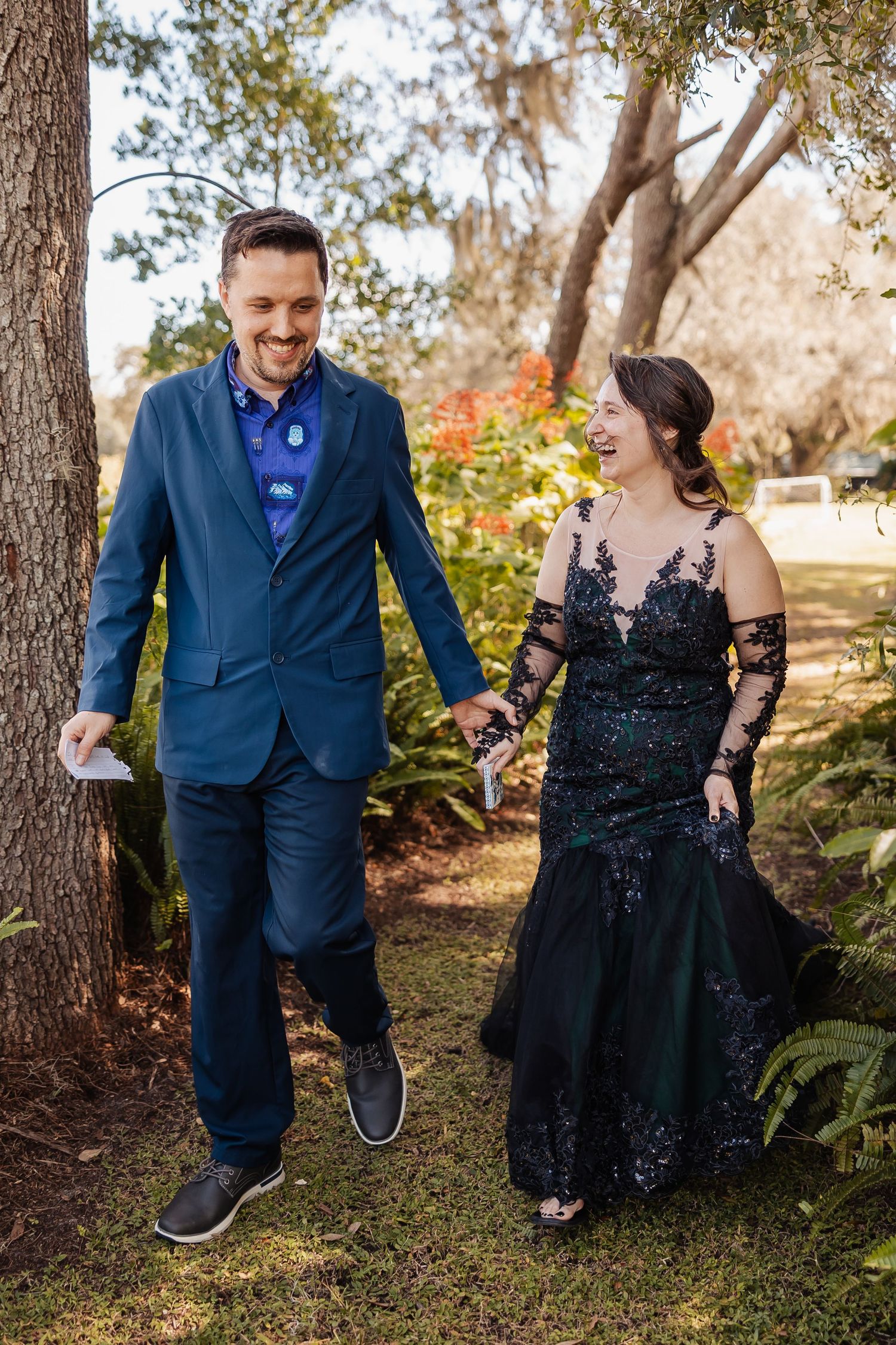 Happy couple walking hand in hand through garden, bride in black lace gown and groom in blue suit.