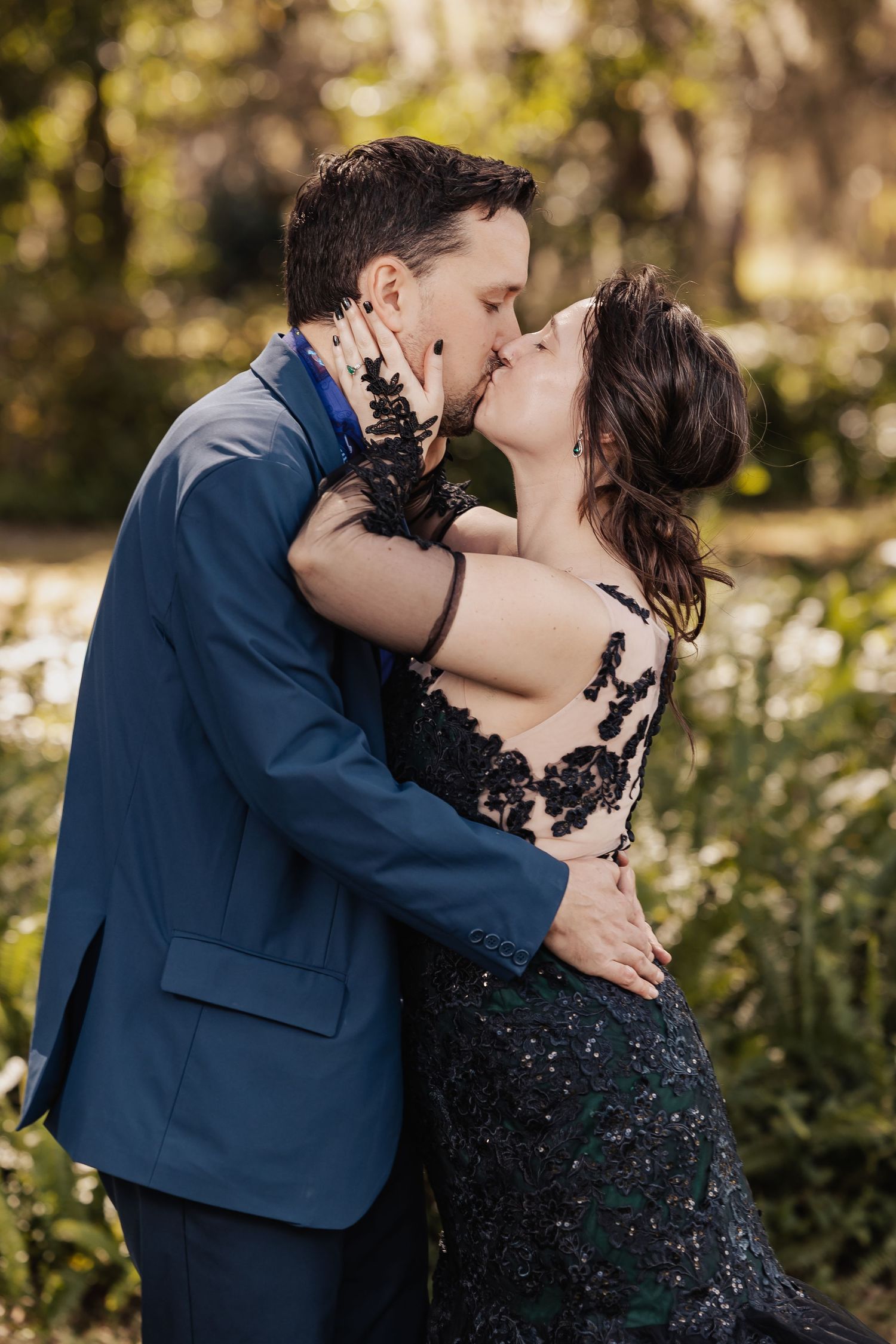 Couple shares passionate kiss surrounded by lush greenery, bride in black lace sequin gown at outdoor wedding.