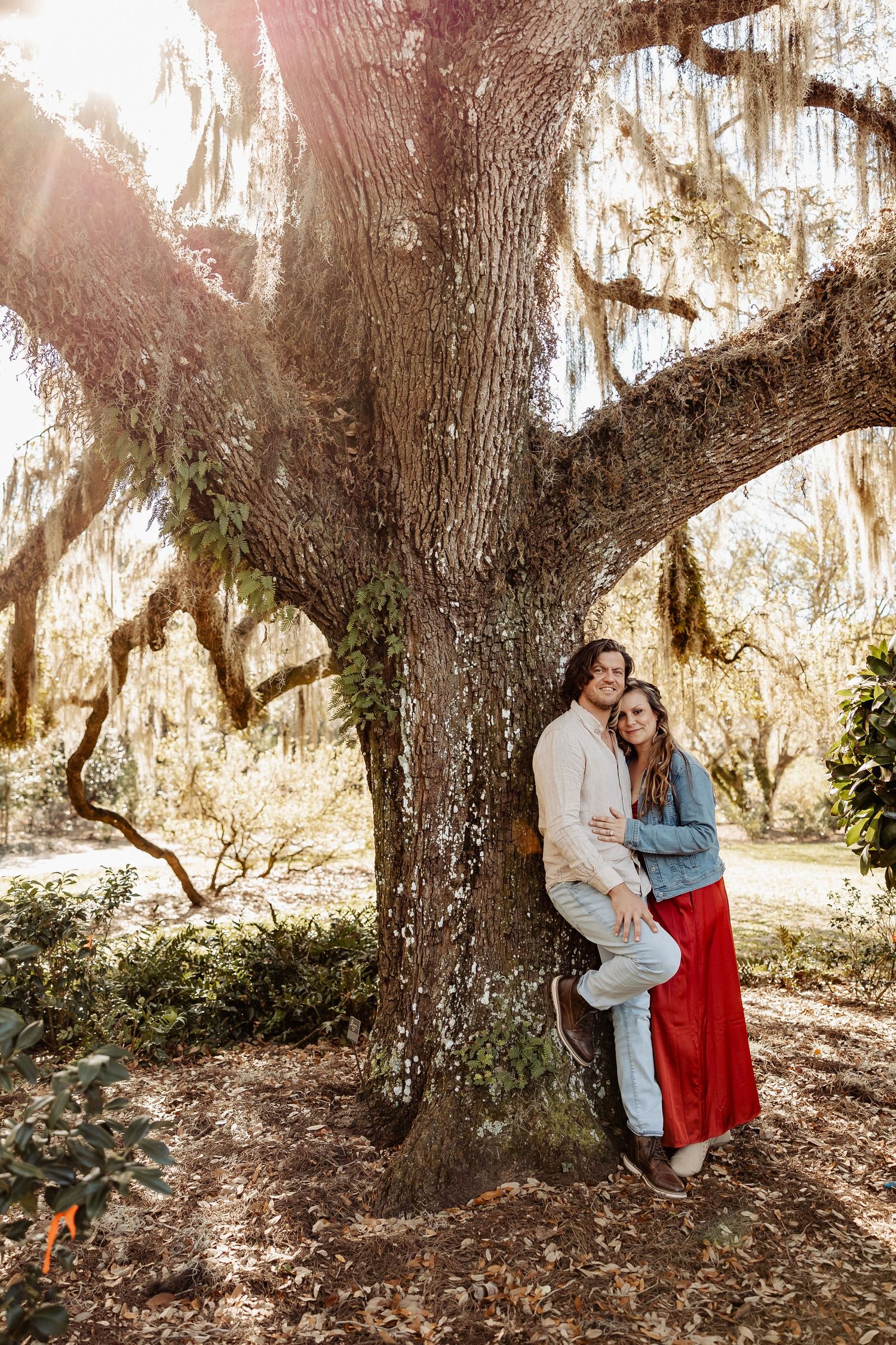 Couple leaning against a large moss-covered oak tree with Spanish moss in a sunlit park.