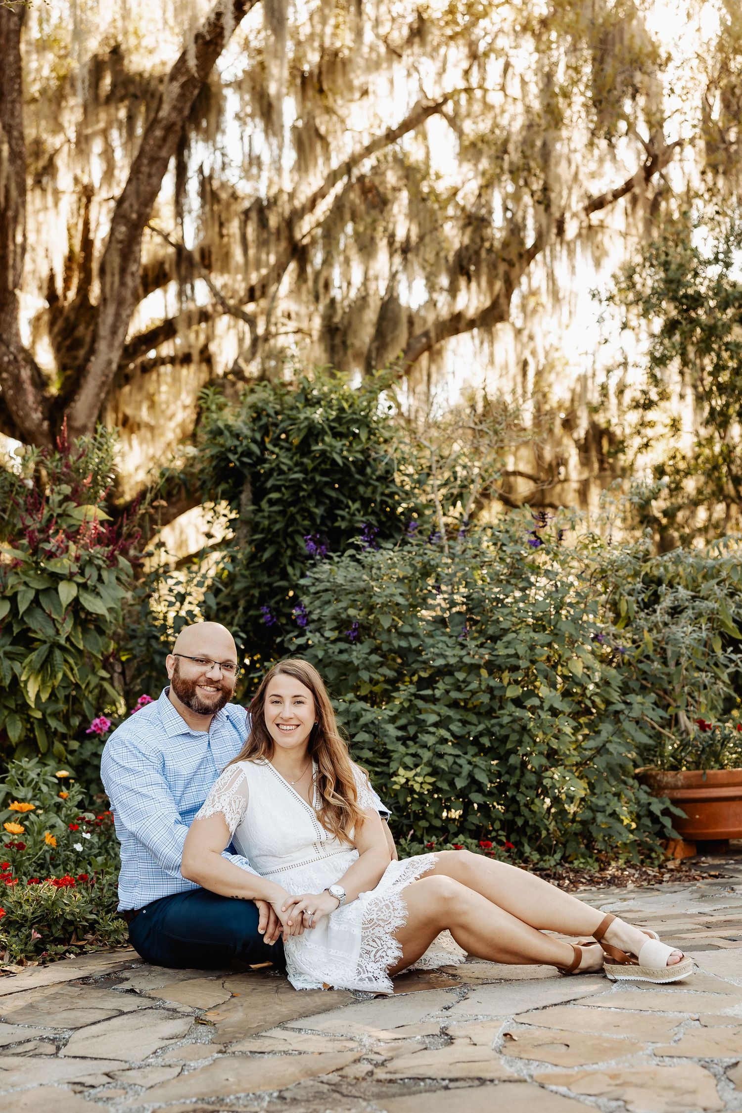 Couple sitting on stone path under sprawling Spanish moss-draped oak tree in a beautiful garden.