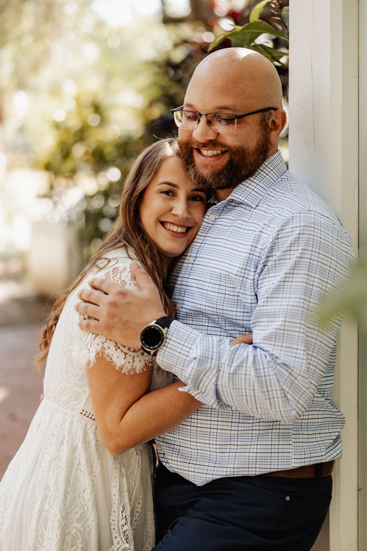 Woman in lace dress hugging smiling bearded man in blue plaid shirt during outdoor engagement photos.