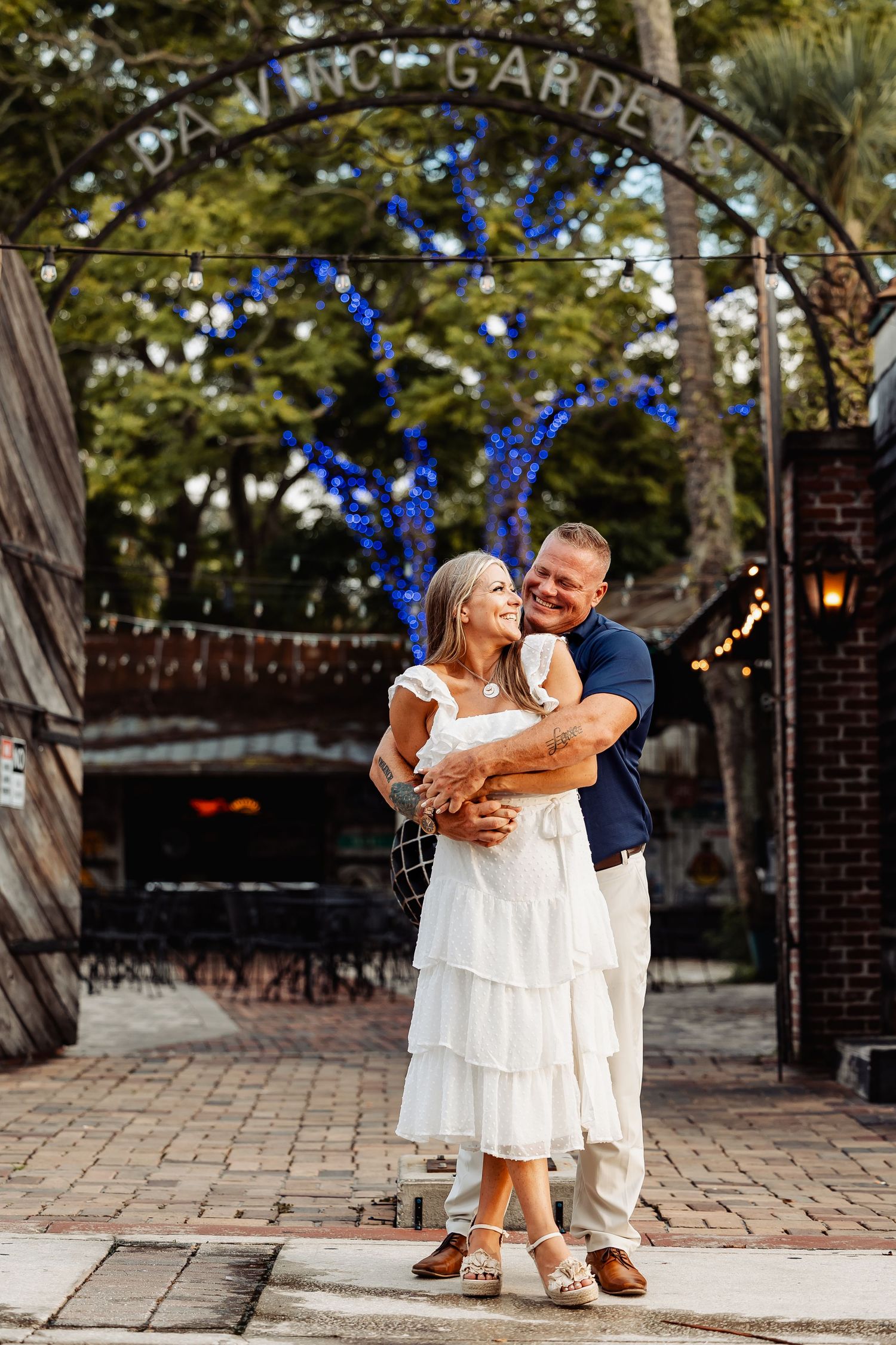 Couple embracing and laughing under a garden gate entrance with blue lit trees in the background.
