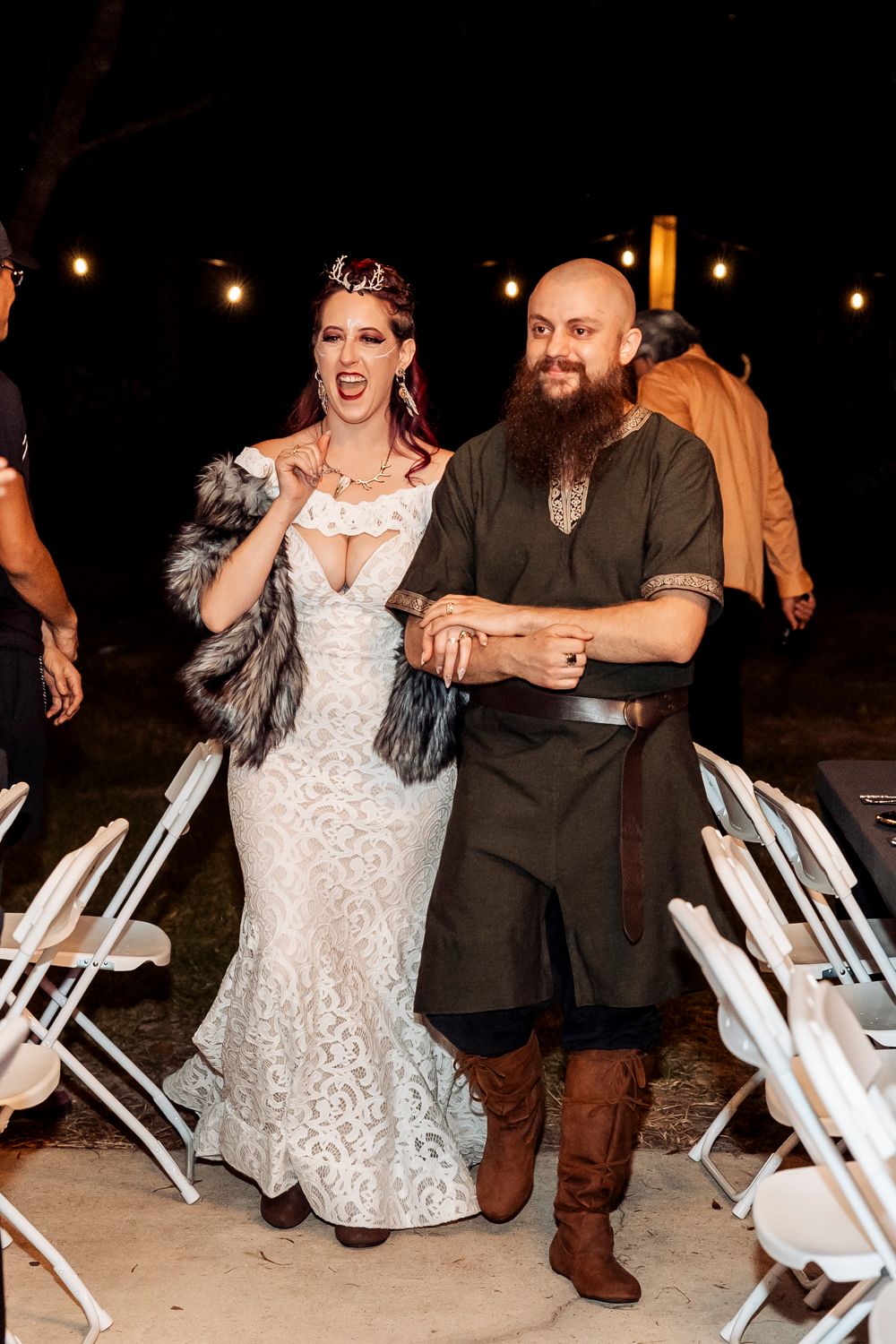 An outdoor wedding reception features medieval-inspired attire and white folding chairs under string lights.
