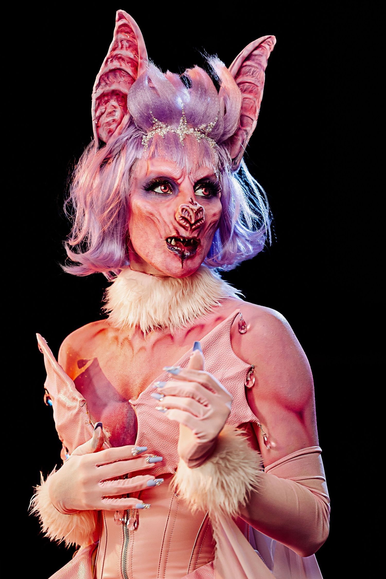 Artistic performer in pink corset and furry wolf-like costume ears poses theatrically against dark background.