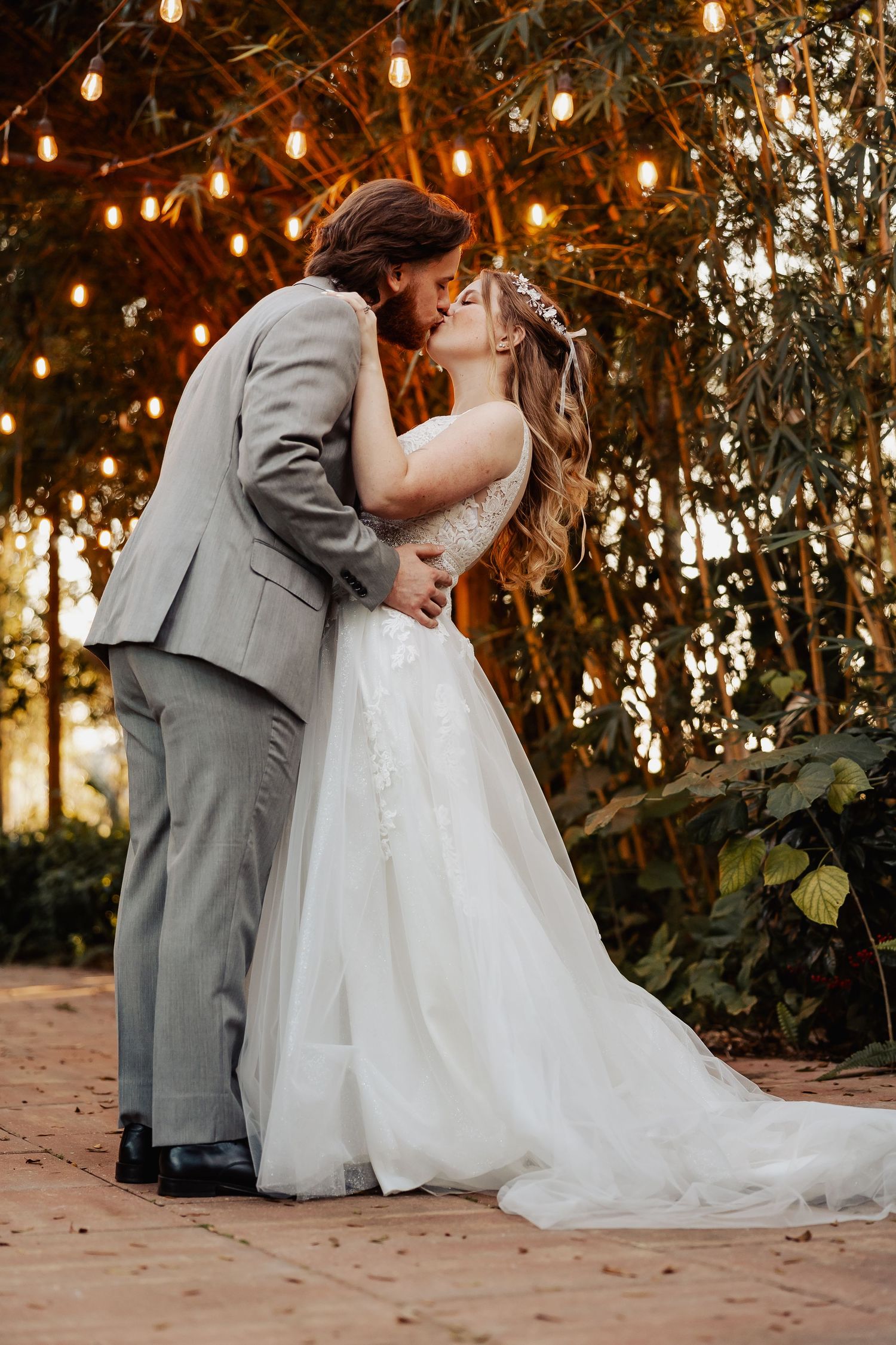 Romantic newlyweds share a kiss under glowing string lights at sunset during their outdoor wedding ceremony.