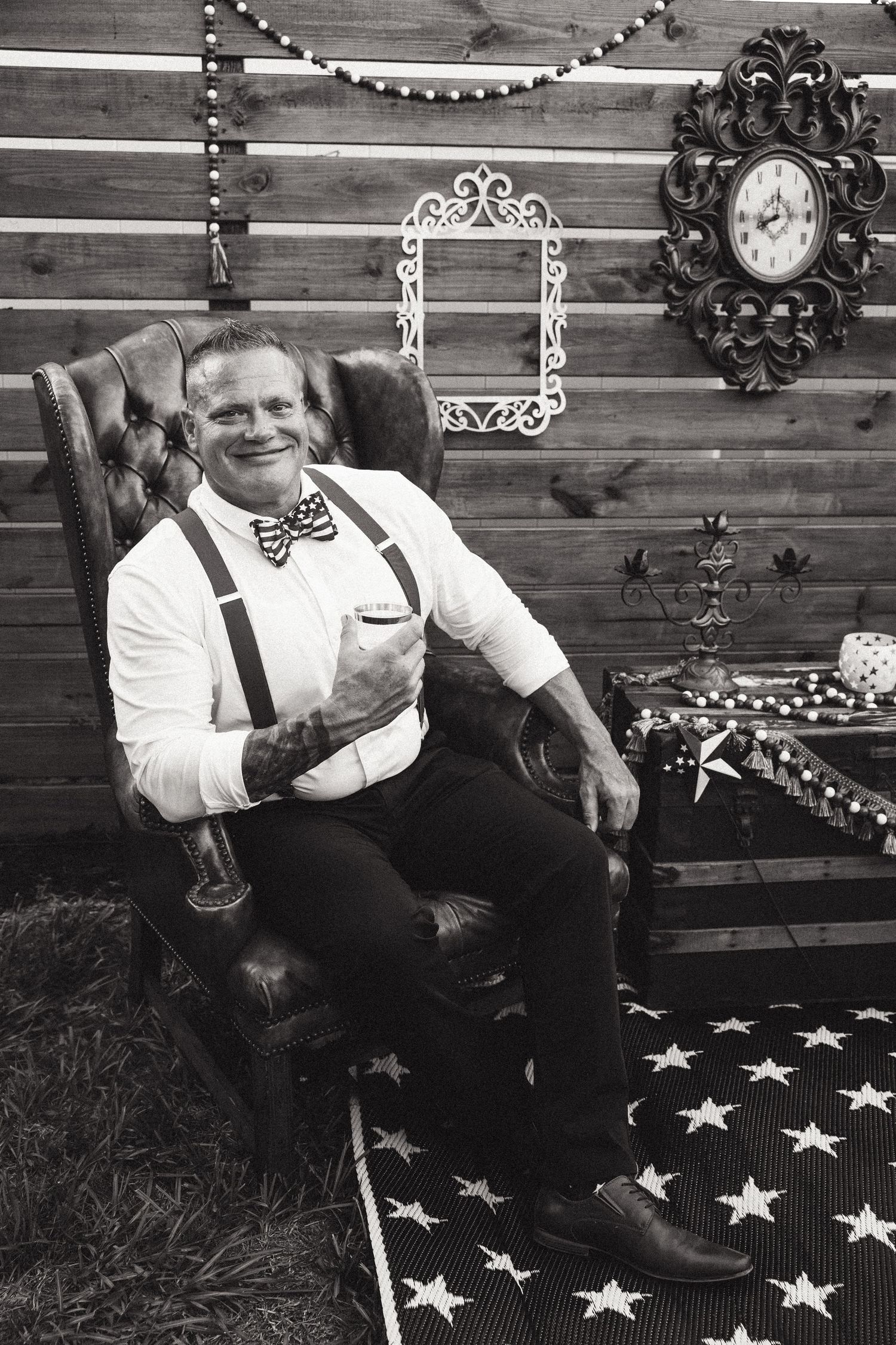 Groom in white shirt and suspenders sitting in leather chair smiling at rustic barn wedding venue.