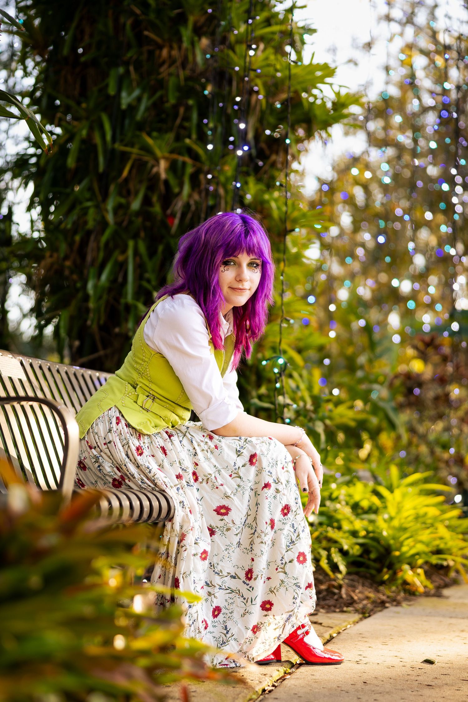 Woman with purple hair in floral skirt and green vest sitting on a park bench surrounded by greenery.