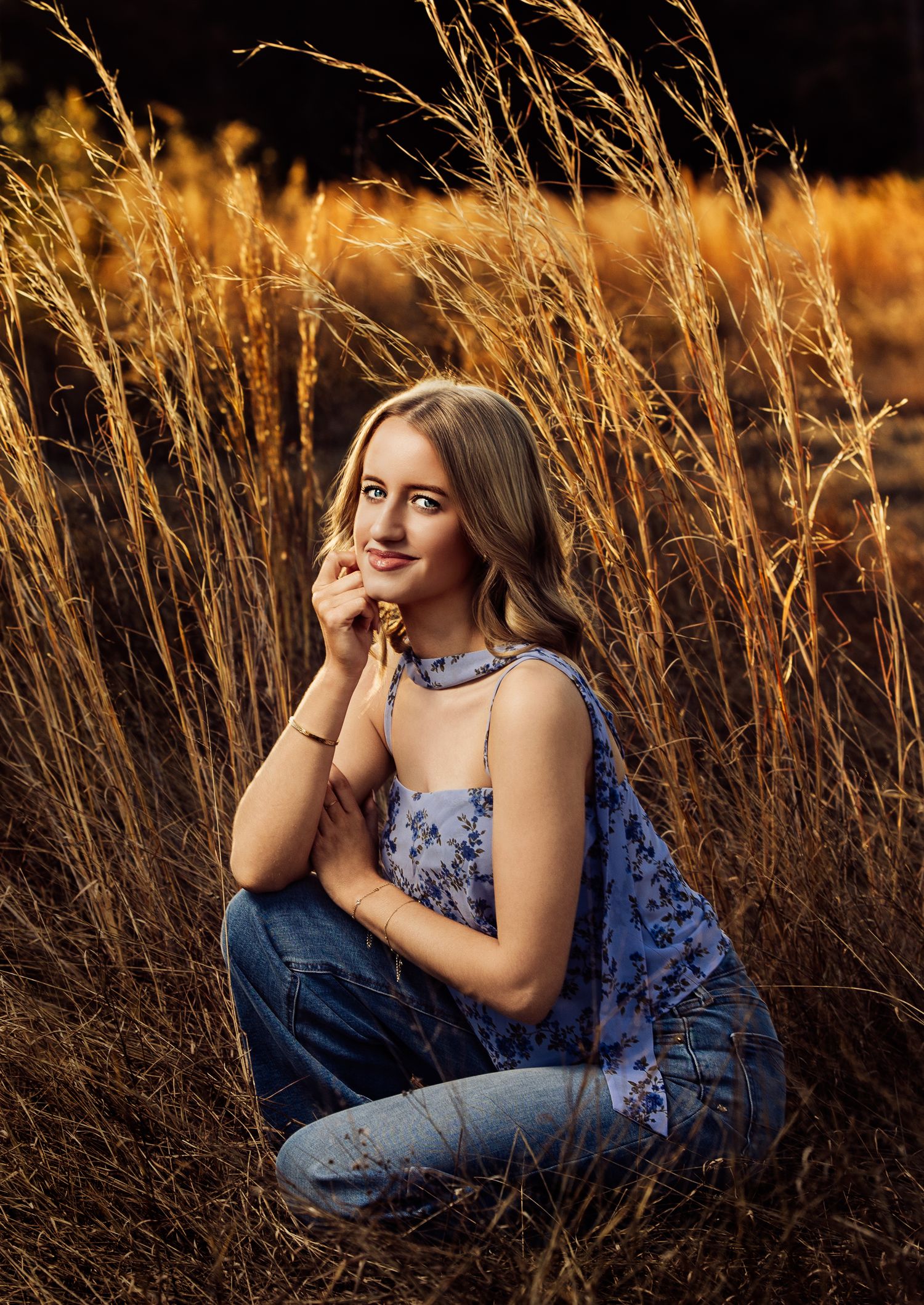 Blonde teenage girl sitting in golden tall grass field wearing blue floral top and jeans.